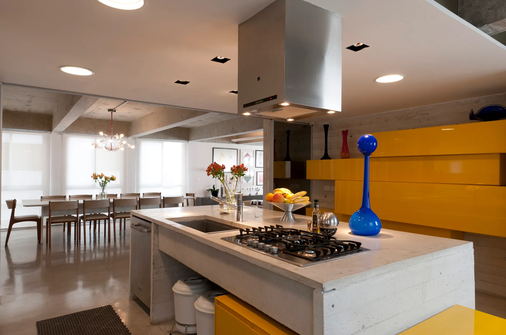 An open kitchen features a concrete island with a built-in cooktop, bright yellow cabinetry, polished concrete floors, and an adjacent dining area.