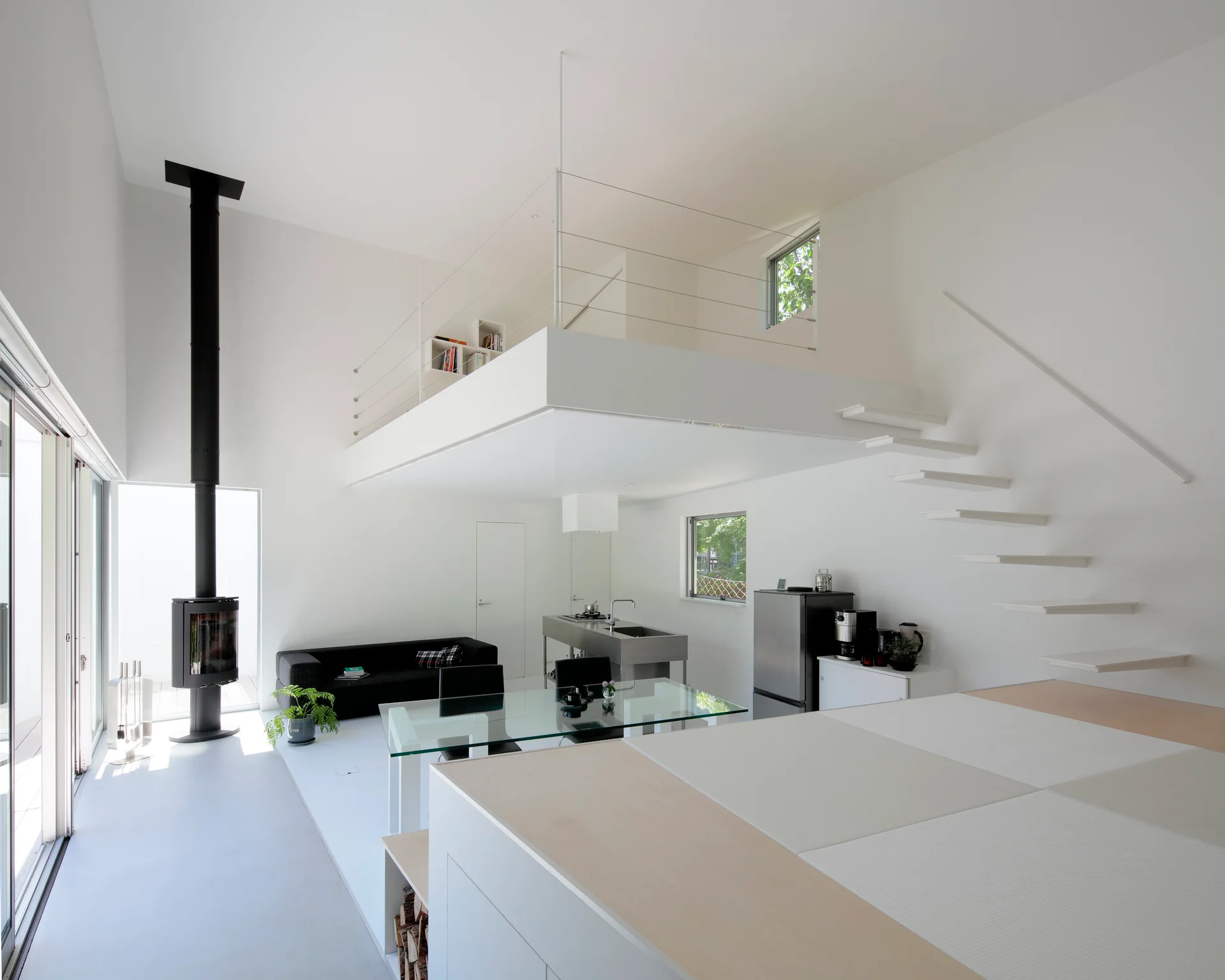 A minimalist double-height white living space featuring a floating staircase, a mezzanine loft, and a tall black freestanding fireplace.