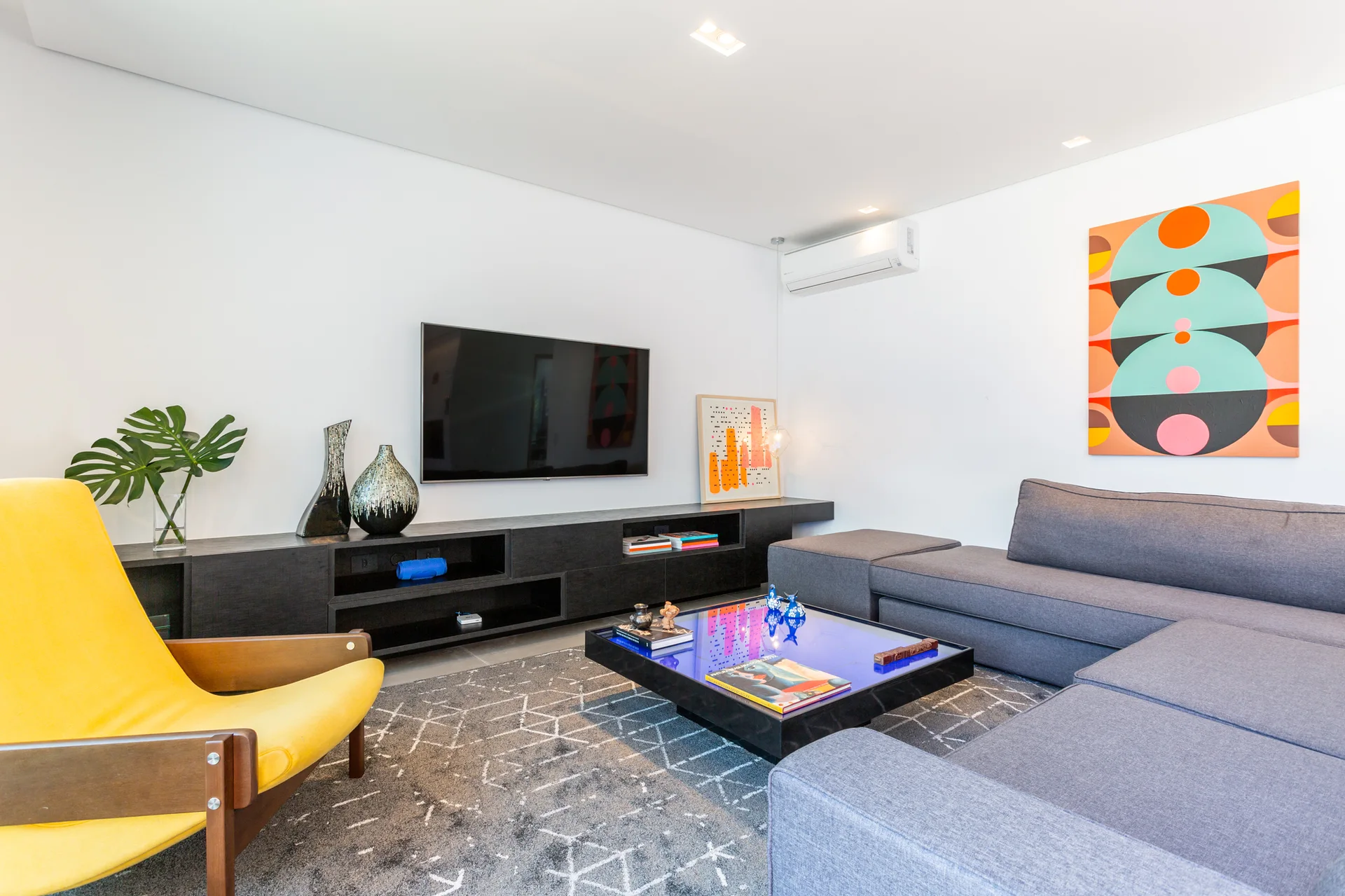 A modern white living room with a grey sectional sofa, a yellow mid-century modern armchair, and a blue glass-topped coffee table.
