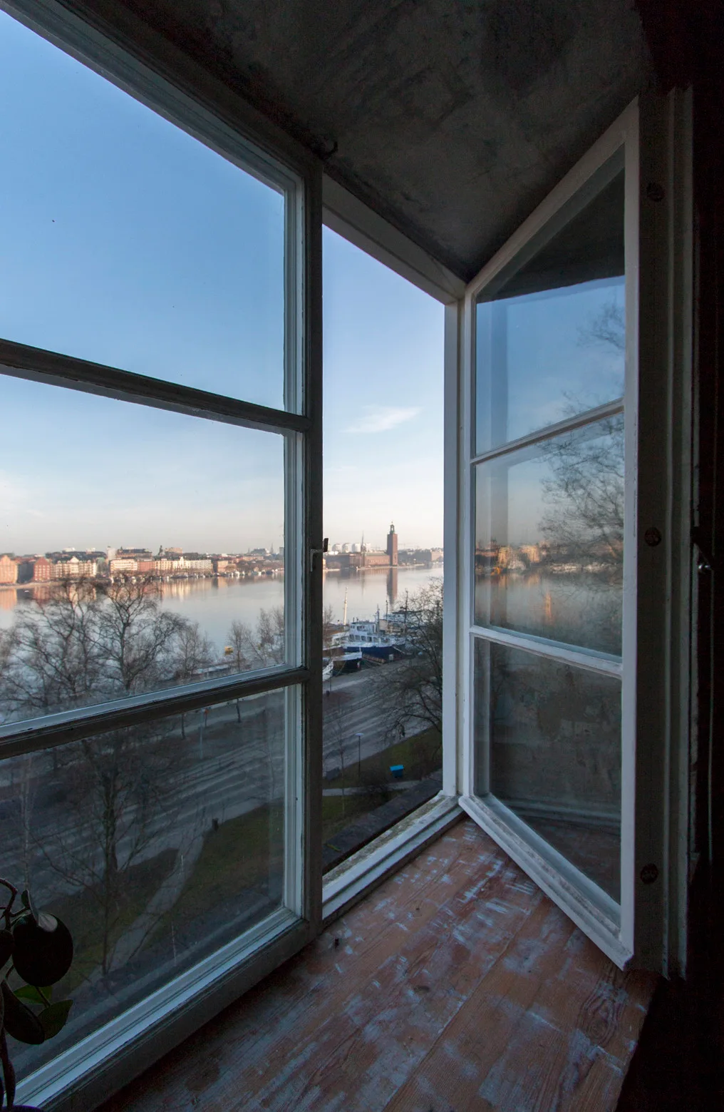 An open white wooden casement window with a wide rustic wood sill looks out over a calm river and distant city skyline.