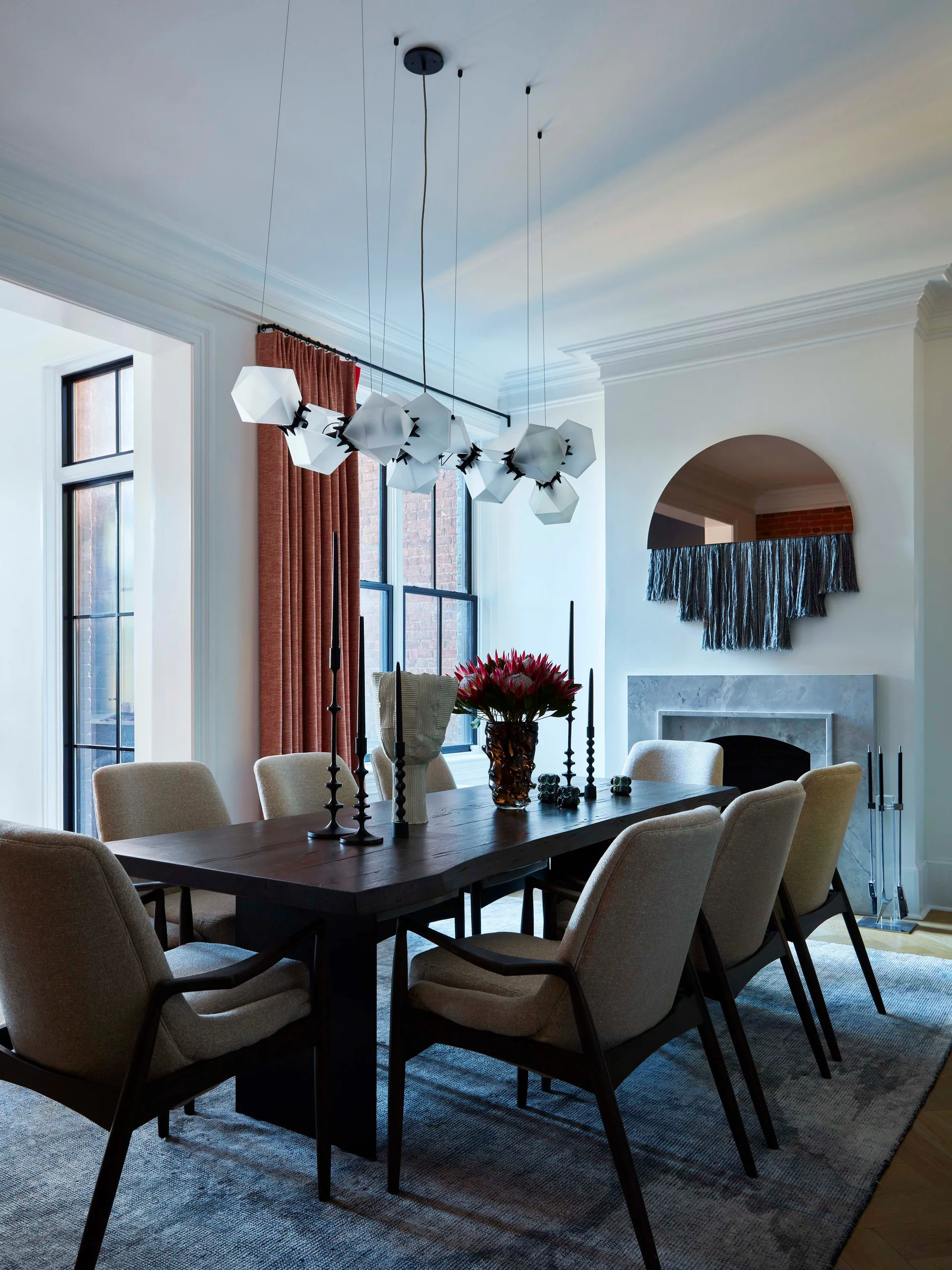 A dining room features a dark wood table with beige upholstered chairs, a geometric white chandelier, and a marble fireplace with an arched mirror.