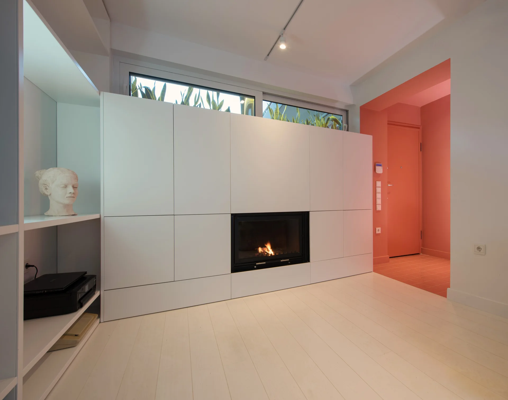 Minimalist white storage cabinets with an integrated black fireplace sit next to a vibrant coral-painted entryway and hallway.