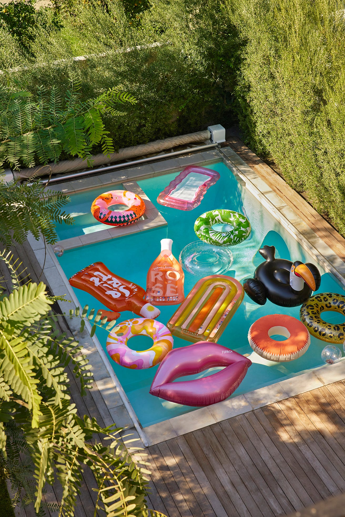 A rectangular backyard swimming pool filled with colorful inflatable floats is surrounded by a wooden deck and high green hedges.