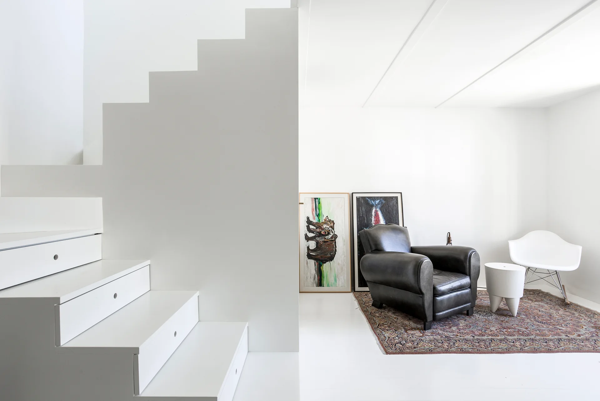 A minimalist white living room features a staircase with built-in storage drawers, a dark leather armchair on a patterned rug, and leaning artwork.