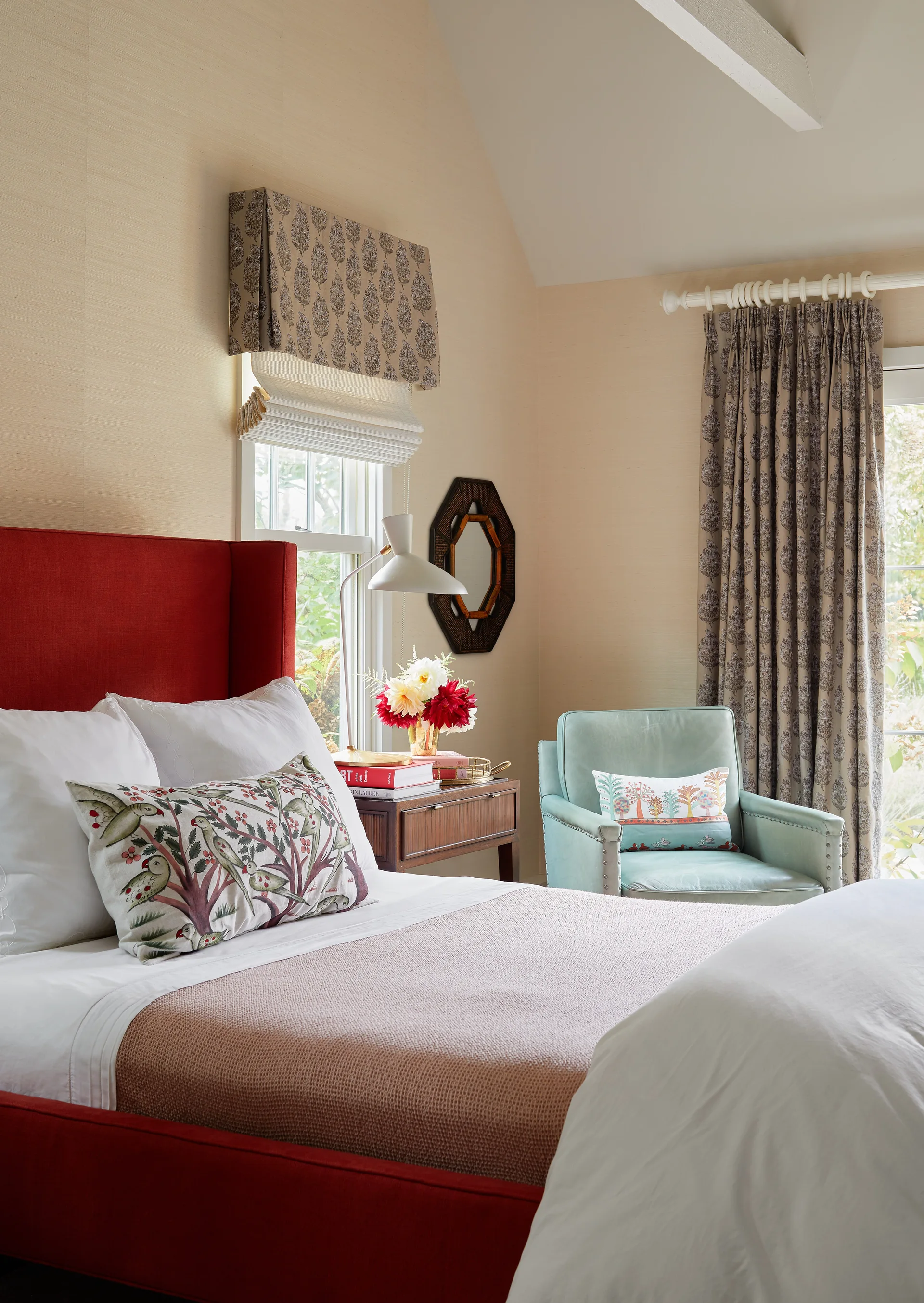 A bedroom featuring a red upholstered headboard, a light blue armchair, grasscloth wallpaper, and botanical patterned curtains.