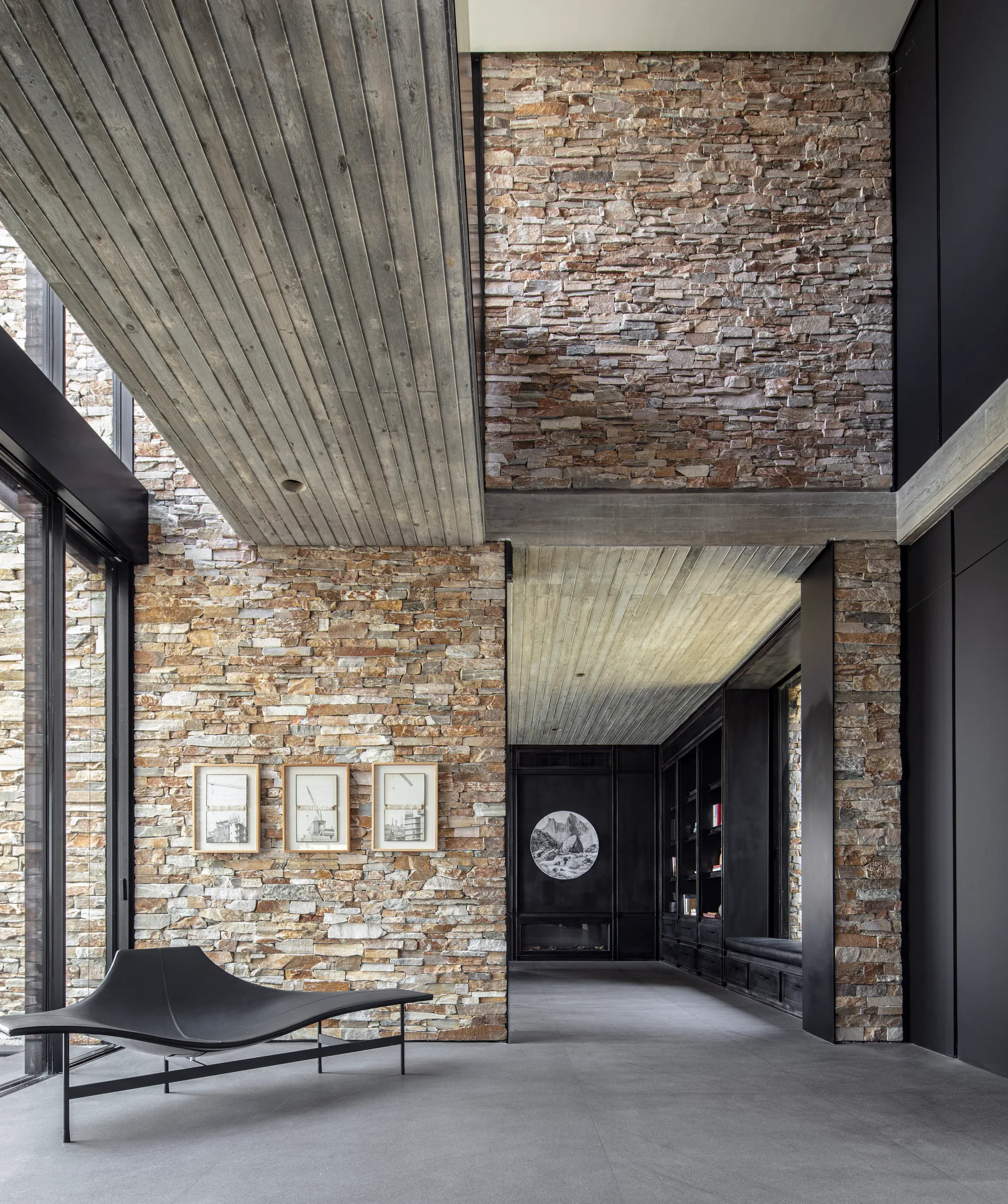 A modern living space featuring rough stacked stone walls, a gray wood plank ceiling, large gray floor tiles, and a minimalist black chaise lounge.