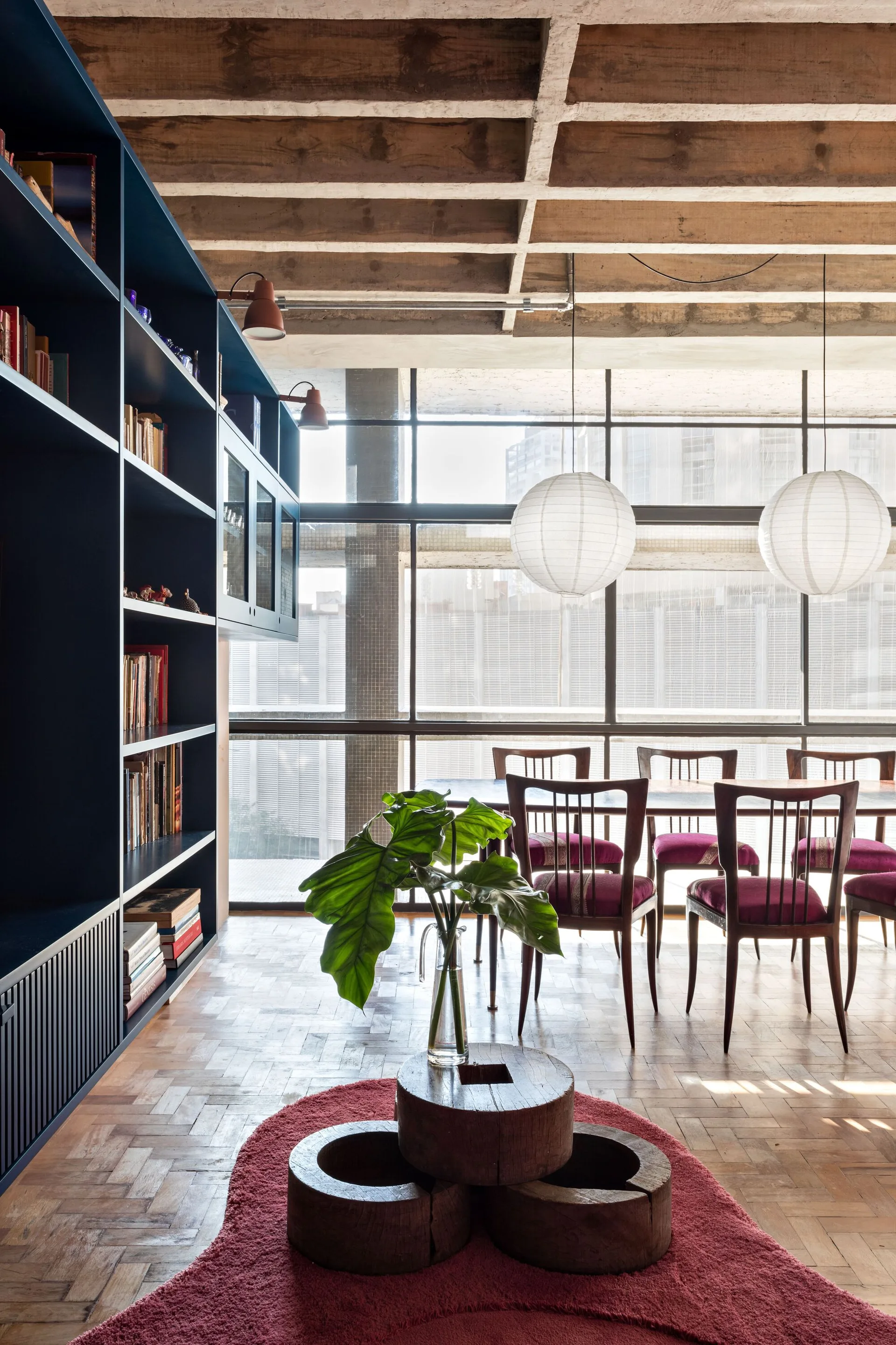 A modern dining room features a wooden table with magenta cushioned chairs, a large navy blue bookcase, and two large spherical paper pendant lights.