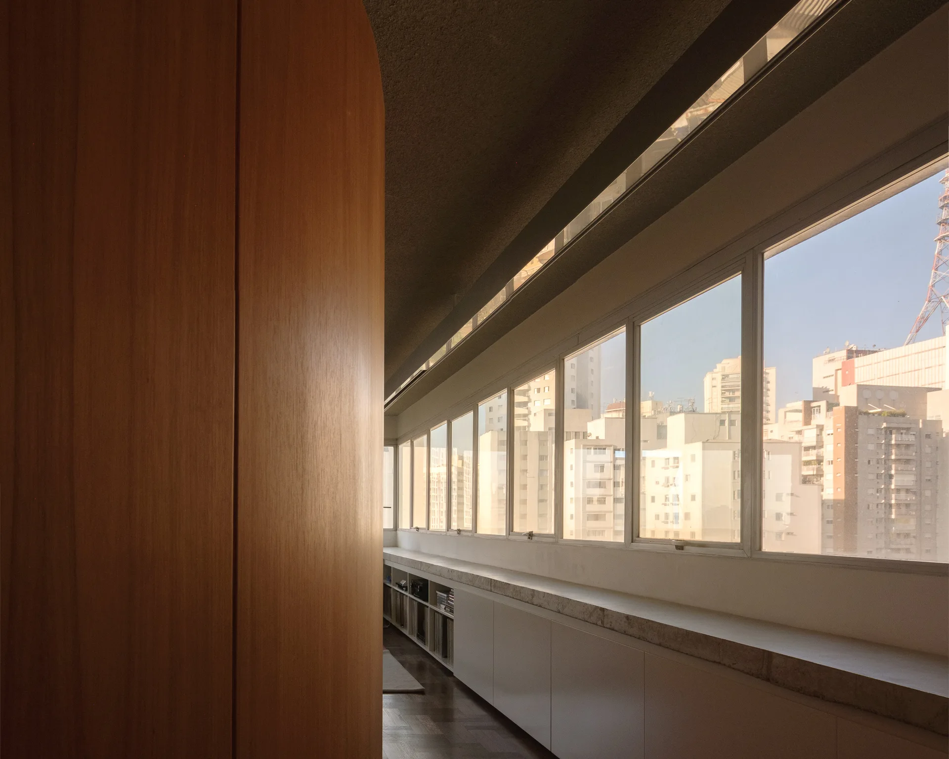 A curved wood partition stands beside a long horizontal window wall featuring low white built-in storage cabinets looking out at city buildings.