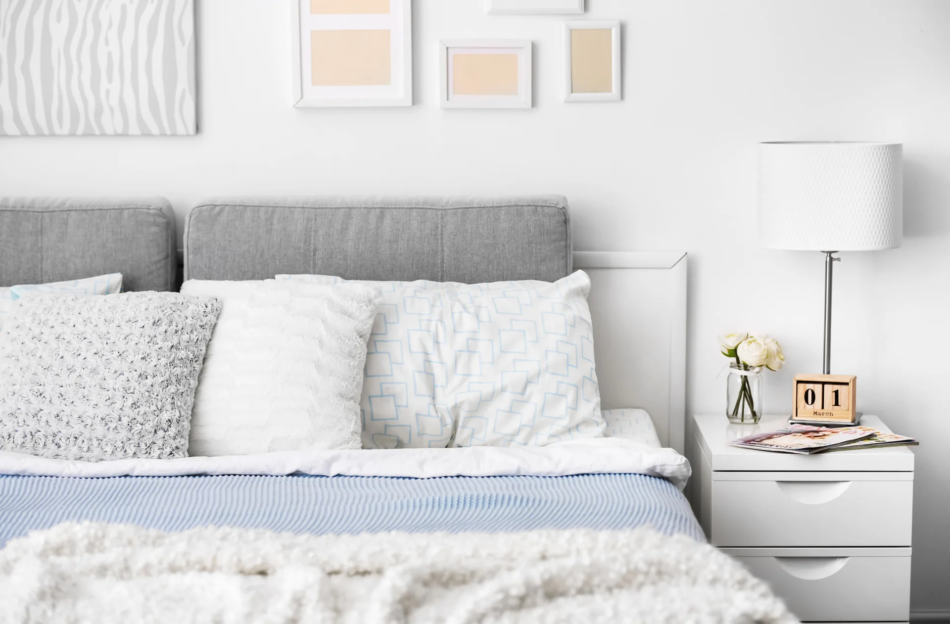 A close-up of a bed with a gray upholstered headboard, white and blue patterned pillows, and a white nightstand holding a white lamp.