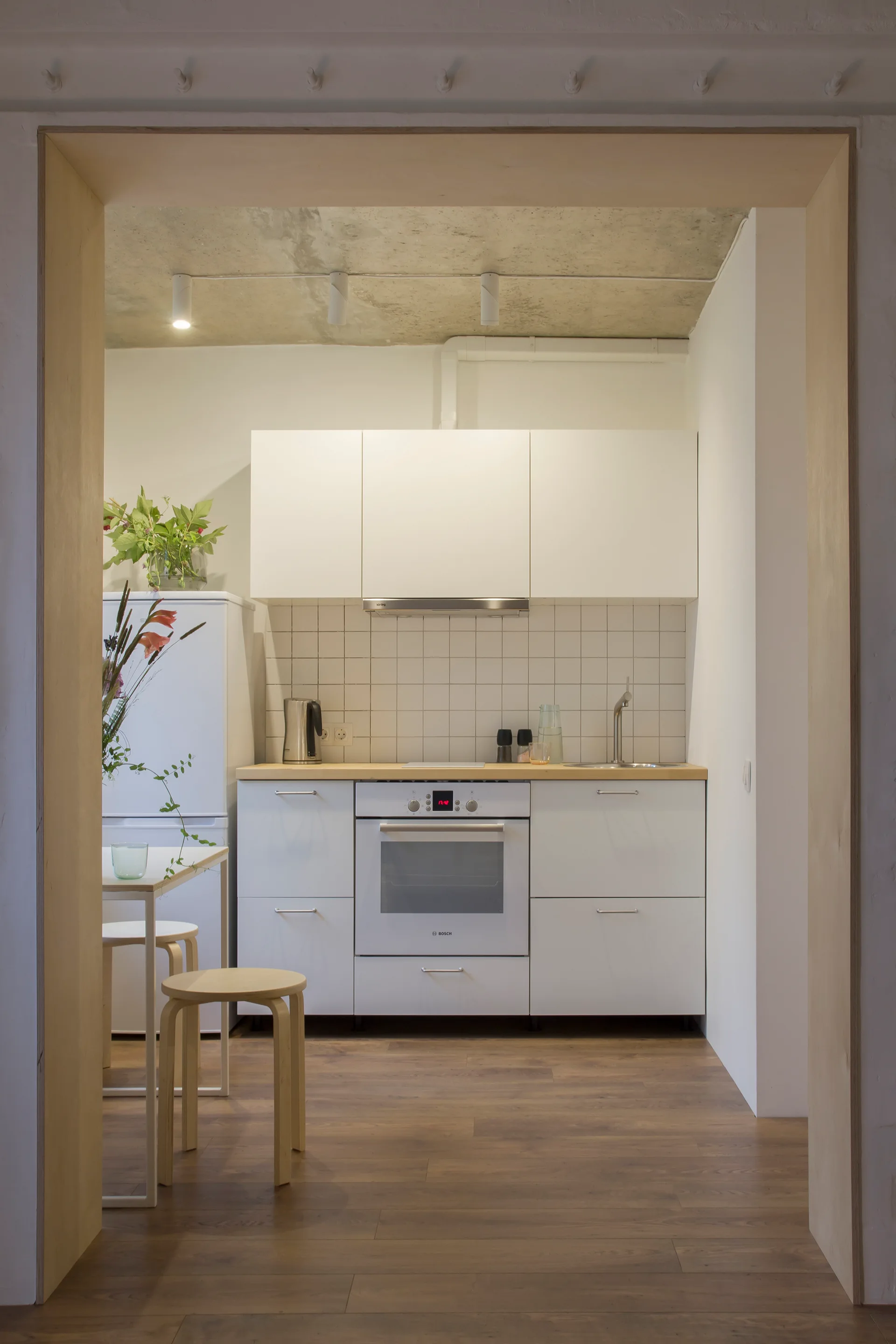 A minimalist kitchen with white cabinets, a wood countertop, and an unfinished concrete ceiling, viewed through a light plywood door frame.