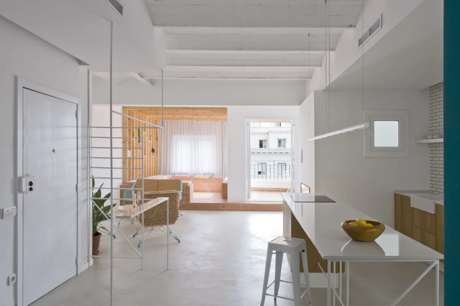 A minimalist open kitchen and living area featuring a white island, exposed ceiling joists, and a raised wooden seating nook in the background.