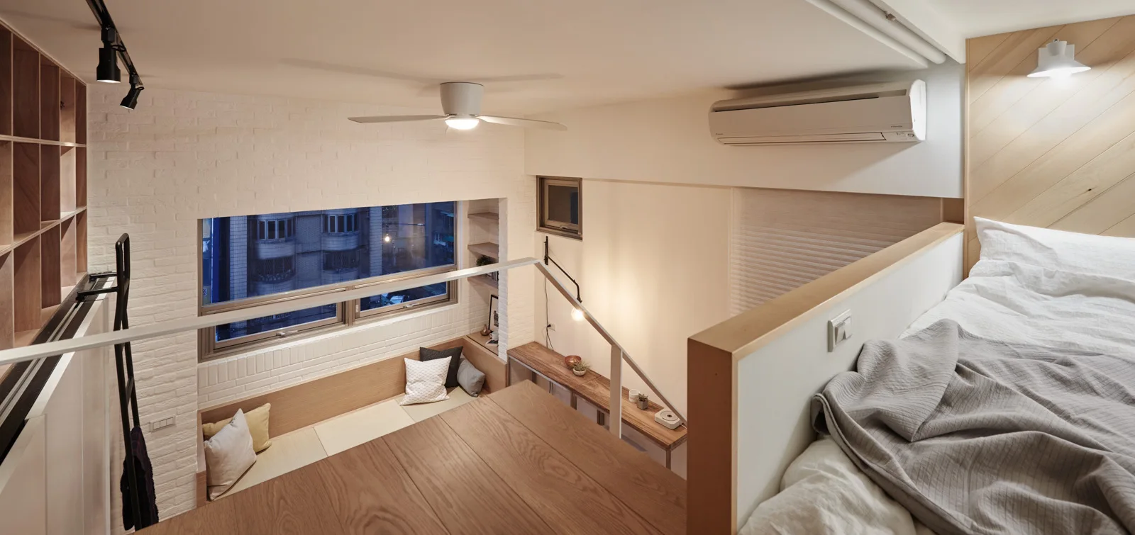 A split-level loft bedroom features a raised wooden platform bed, a built-in window bench with throw pillows, and a white painted brick wall.