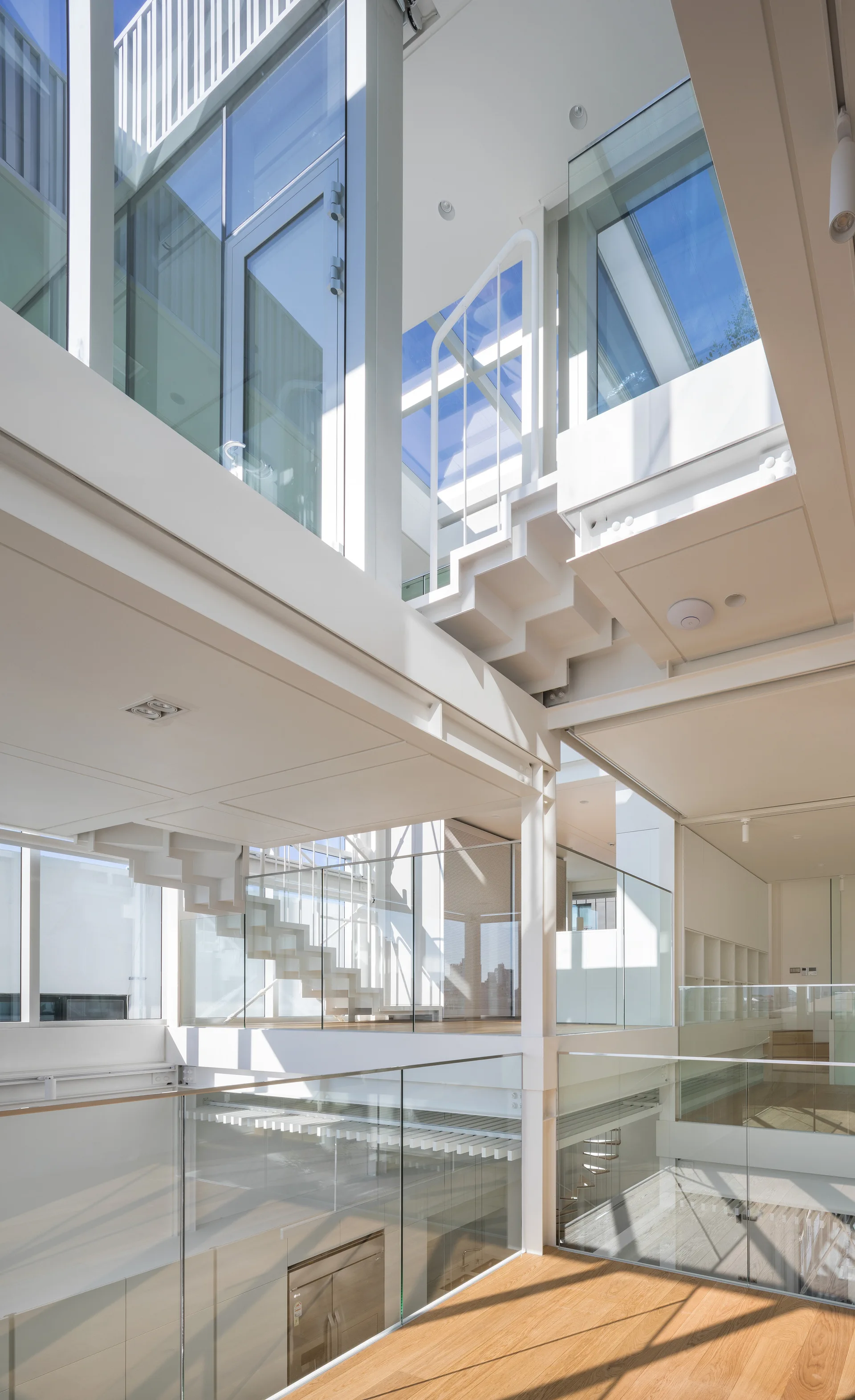 A bright multi-level modern interior with a white steel staircase, transparent glass railings, and natural wood flooring under a skylight.