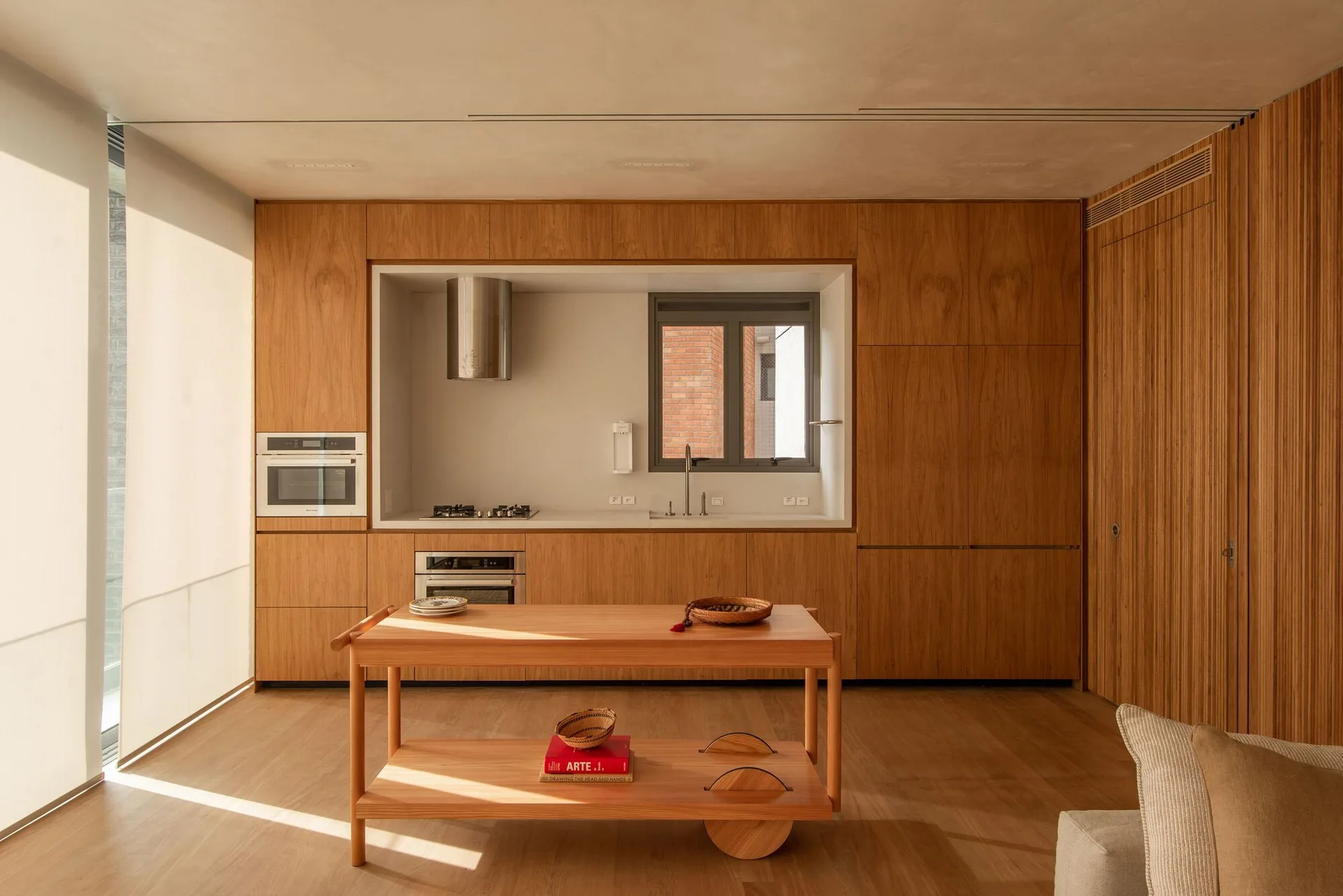 A minimalist kitchen features floor-to-ceiling wood cabinetry, a recessed gray cooking alcove with a sink, and a wooden mobile island on floor planks.