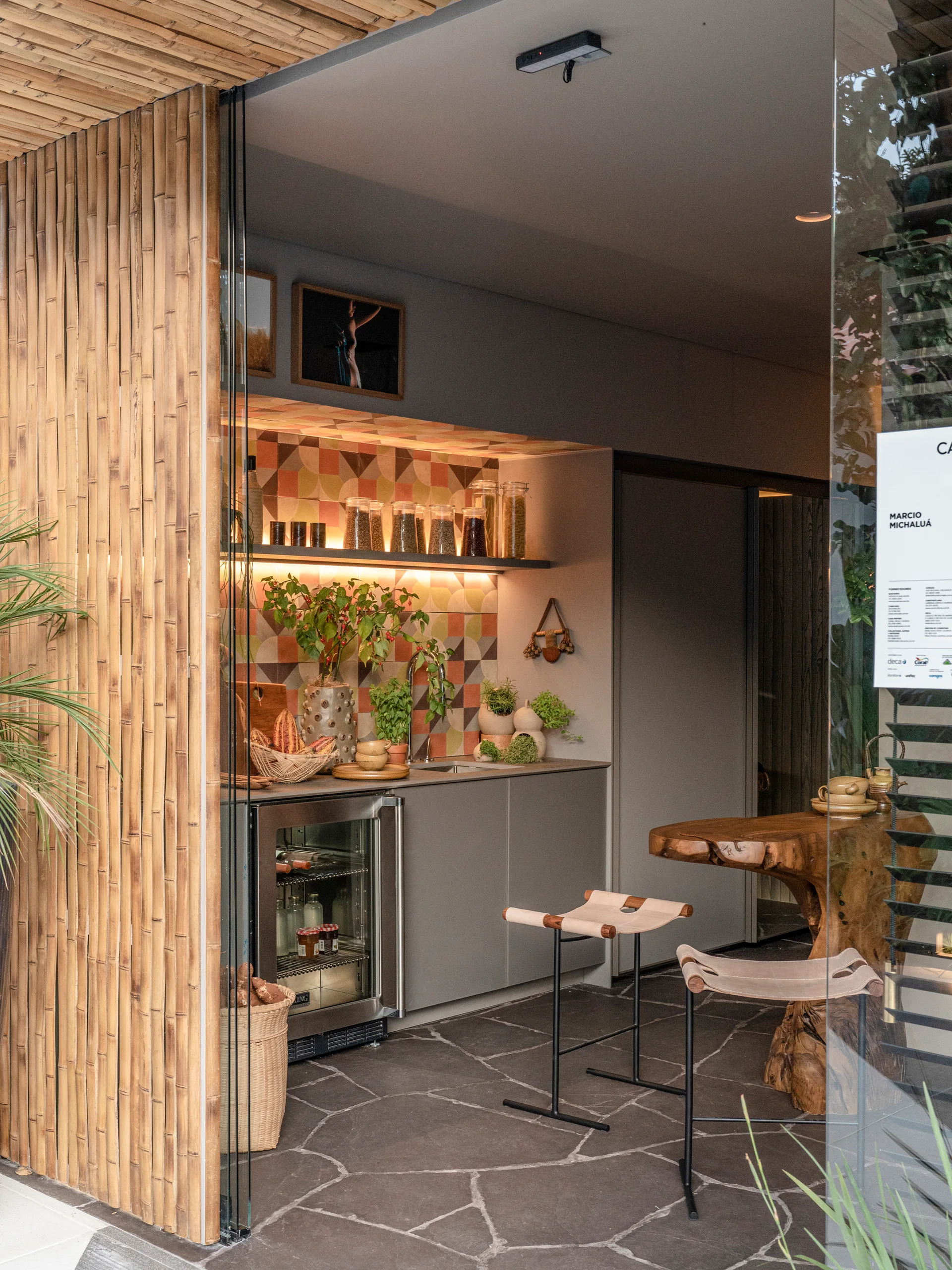 An indoor-outdoor kitchenette features a geometric tile backsplash, gray cabinets, a small beverage fridge, and a live-edge wooden table with stools.