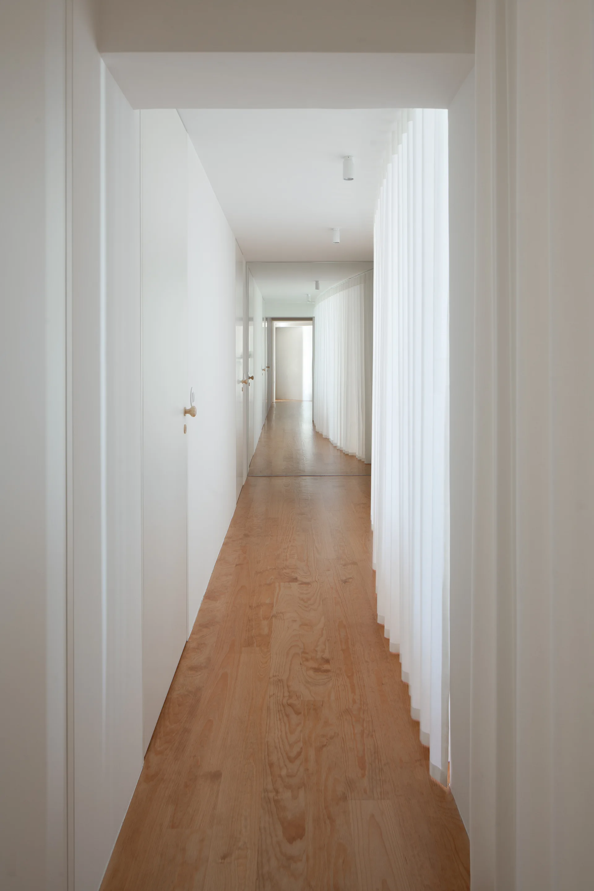A long minimalist corridor with light wood flooring, seamless white flush doors, and a long pleated white fabric curtain hanging from the ceiling.