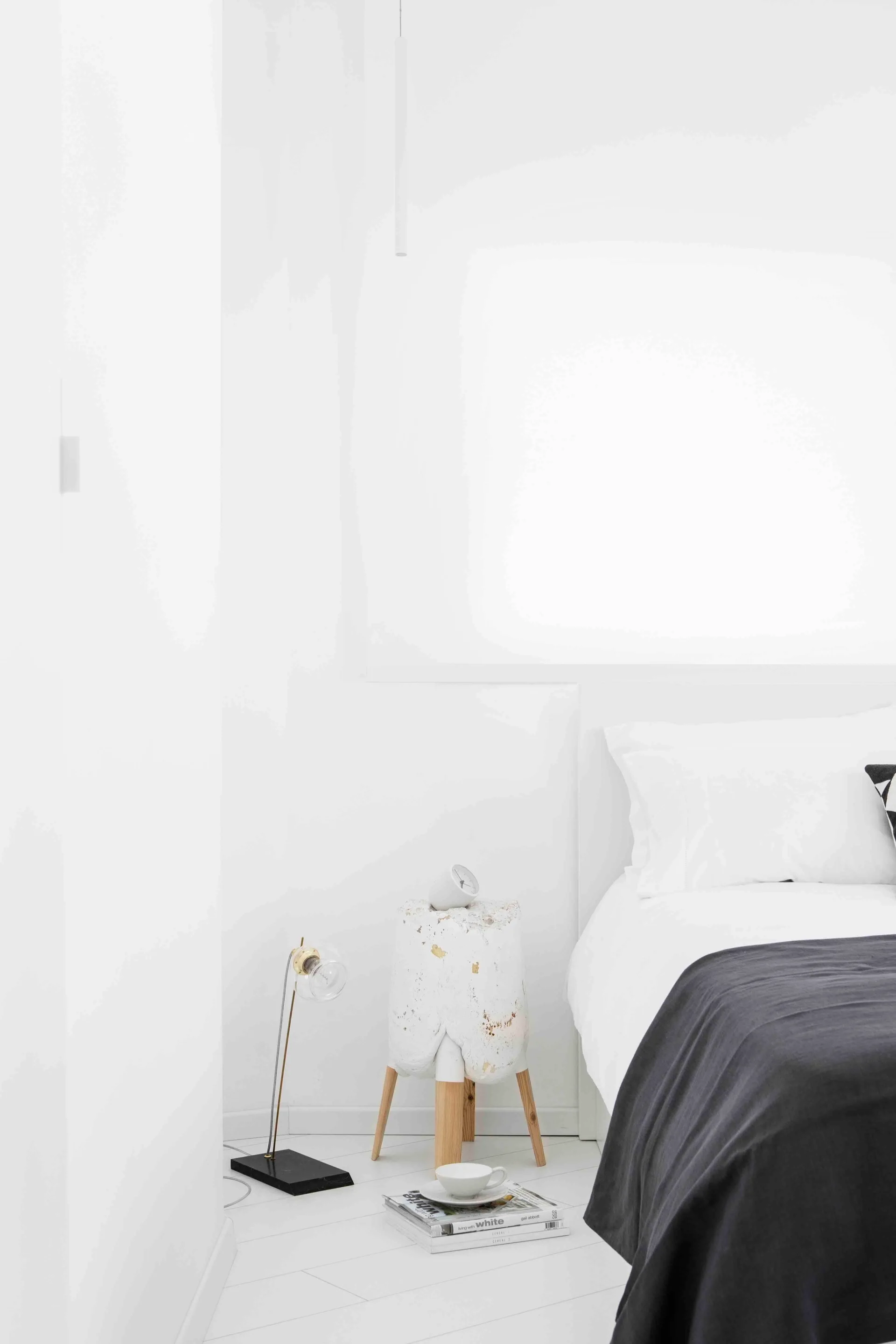 A minimalist bedroom features white walls, a bed with a dark grey throw, and a textured white plaster side table with wooden legs.