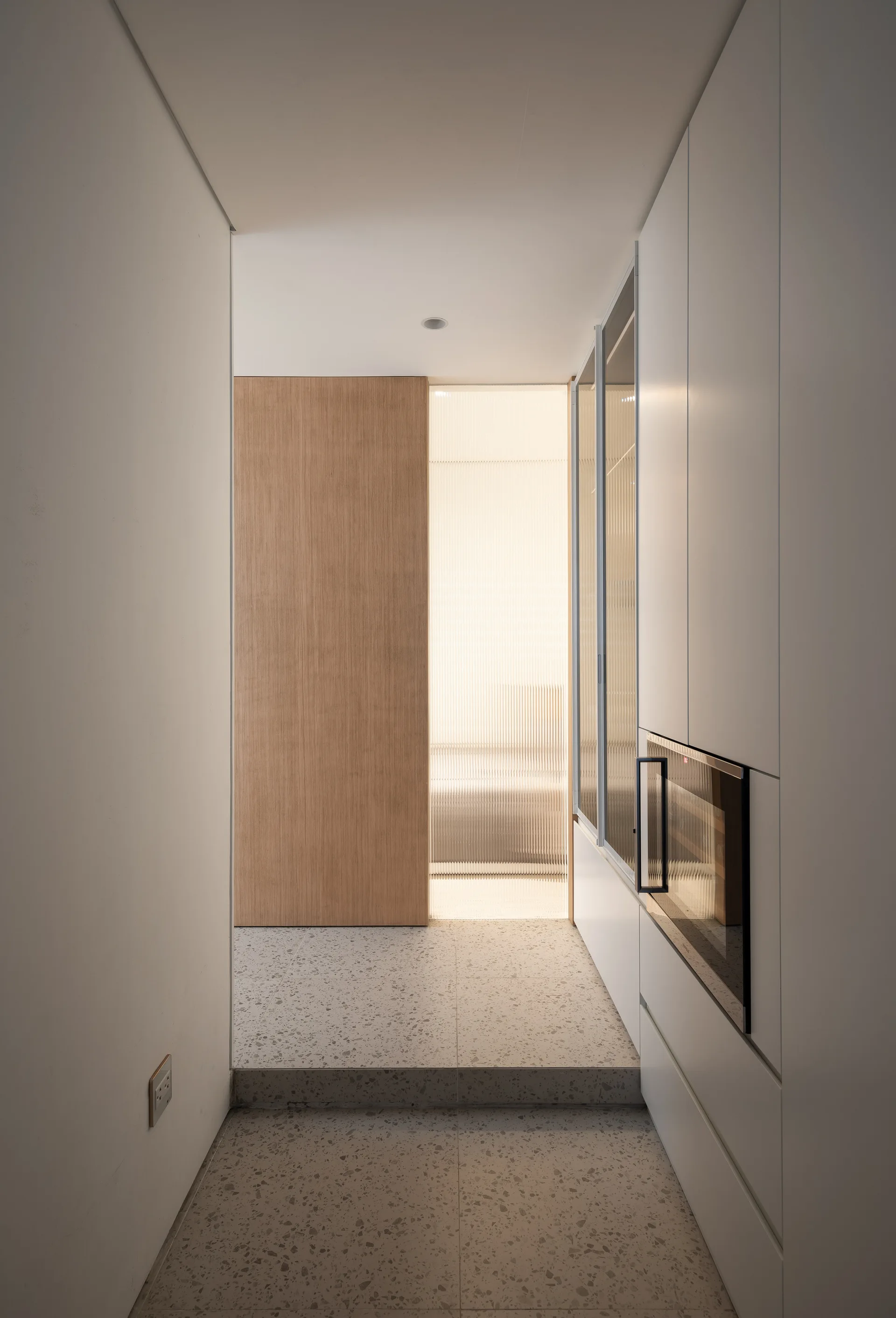 A minimalist hallway featuring seamless white cabinetry, grey terrazzo tiled flooring with a step, and a backlit fluted glass partition next to a wooden door.