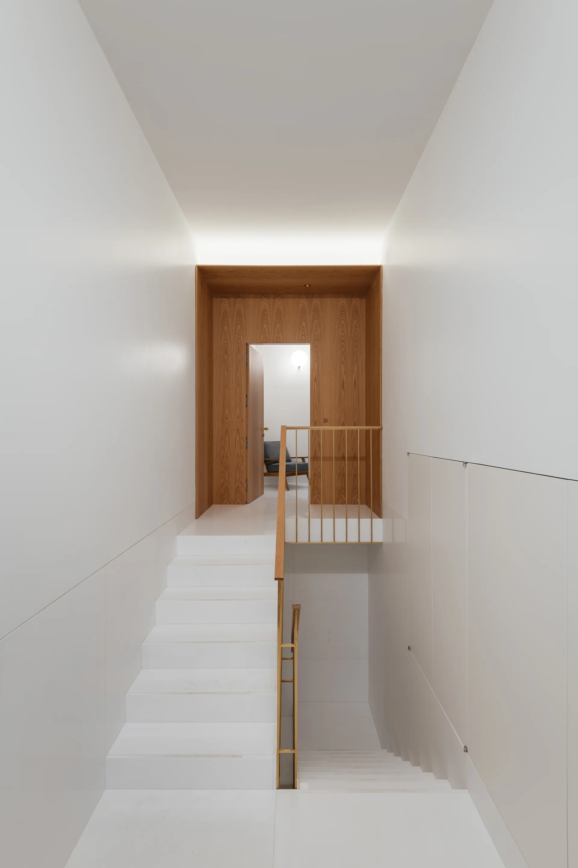 Minimalist white staircase with thin brass railings leading to a landing framed in warm wood paneling with an open doorway.