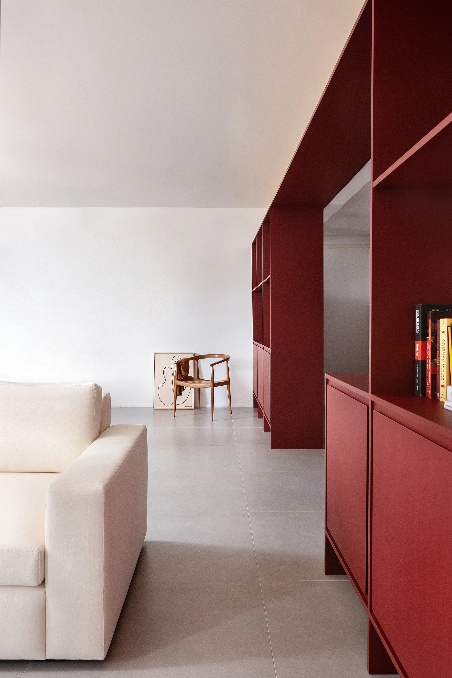 A minimalist living room features a large matte red built-in bookshelf partition, a beige sofa, and a light wood armchair against a white wall.