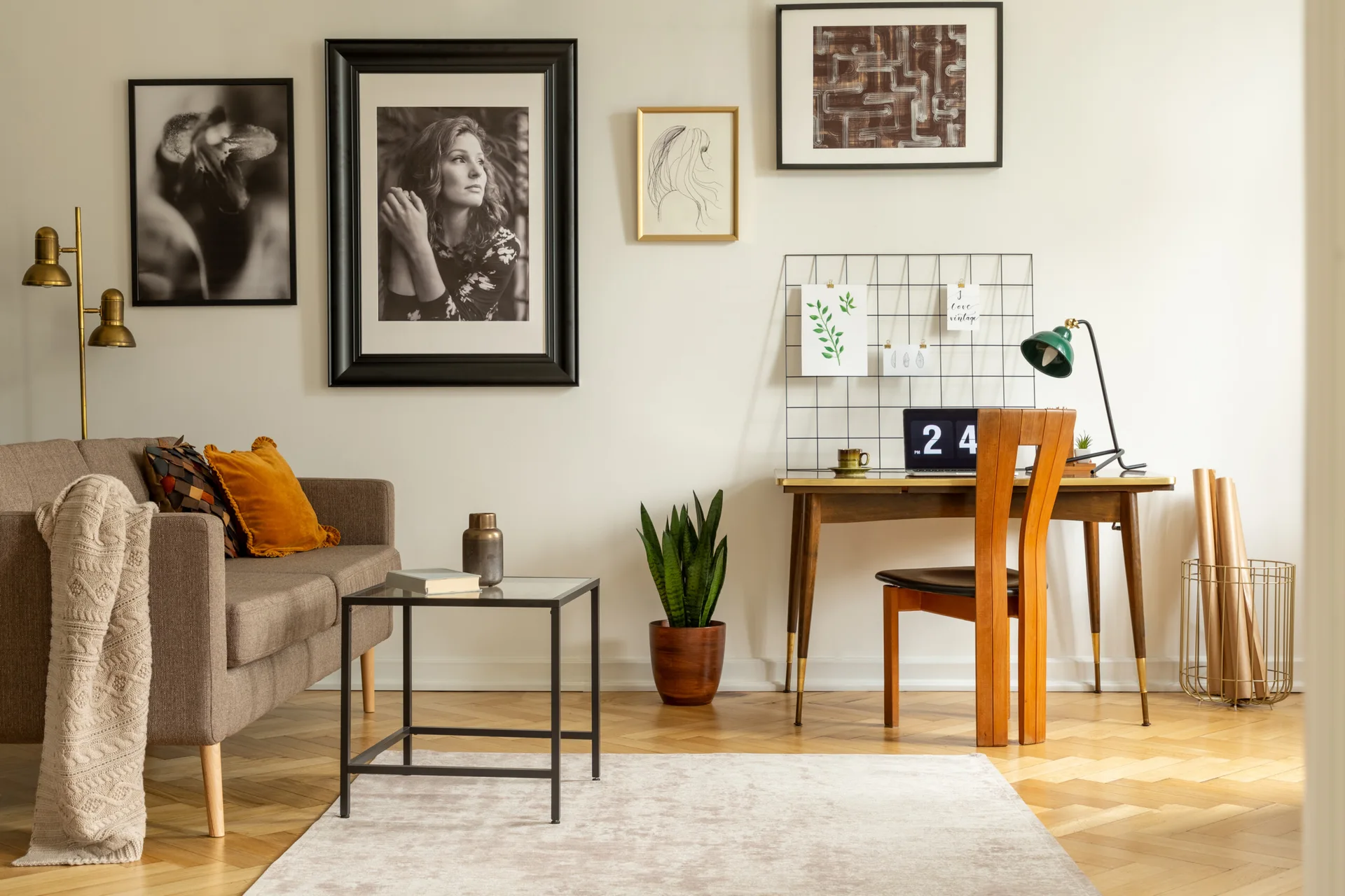 A warm-toned living room features a brown sofa, a wooden writing desk, a snake plant, and framed artwork on the wall above herringbone parquet flooring.