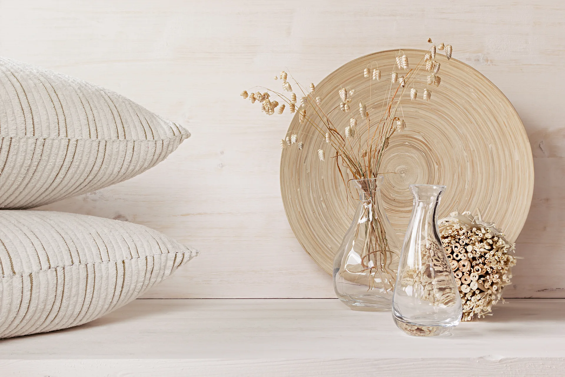 Two stacked cream striped pillows next to glass vases and a round wooden plate against a light wood background.