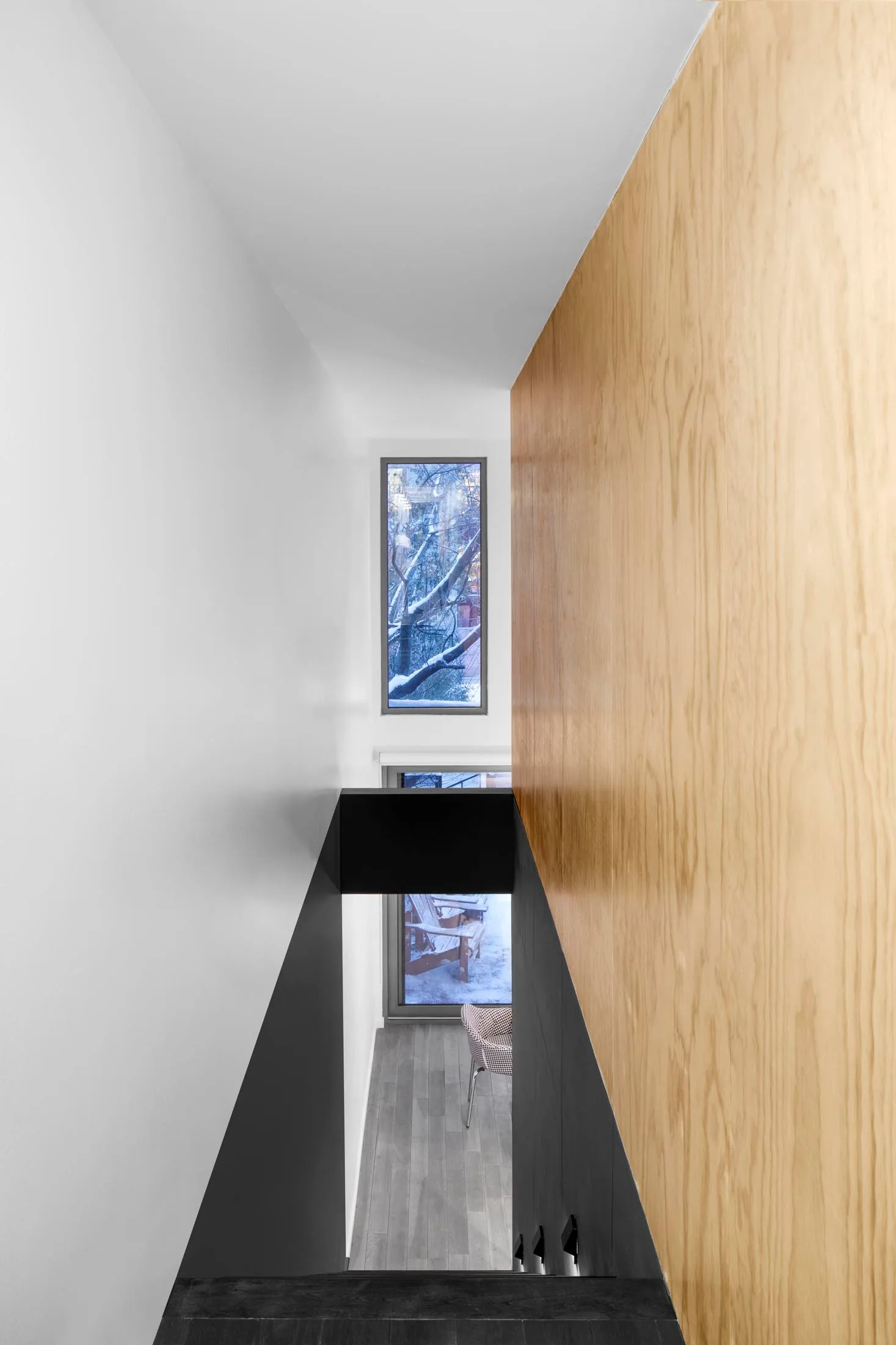 A high-angle view looking down a modern minimalist stairwell with a tall light wood wall, white plaster, and matte black railings.