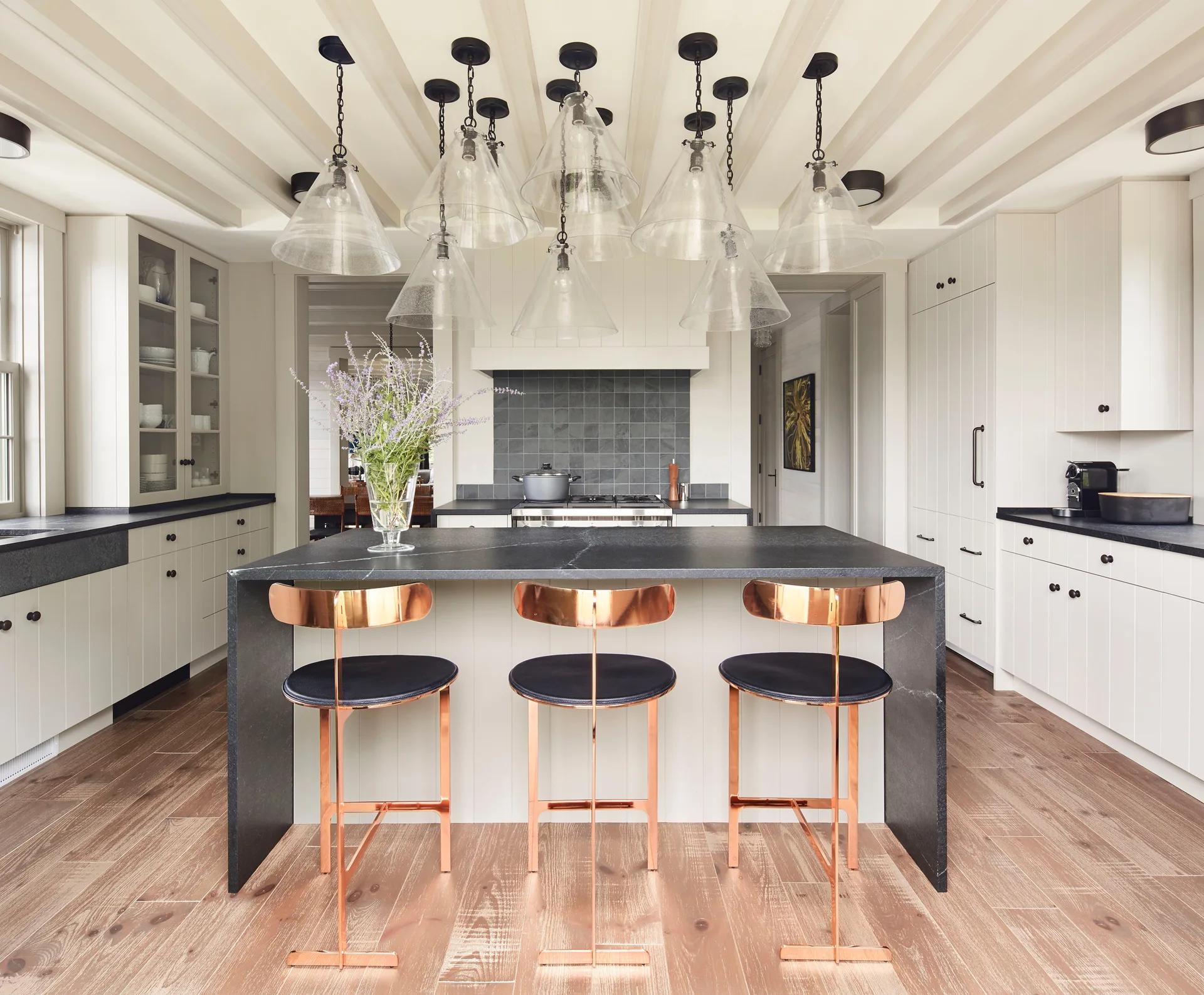 A modern kitchen with a black stone waterfall island, three copper bar stools, cream cabinetry, and clustered glass pendant lights.