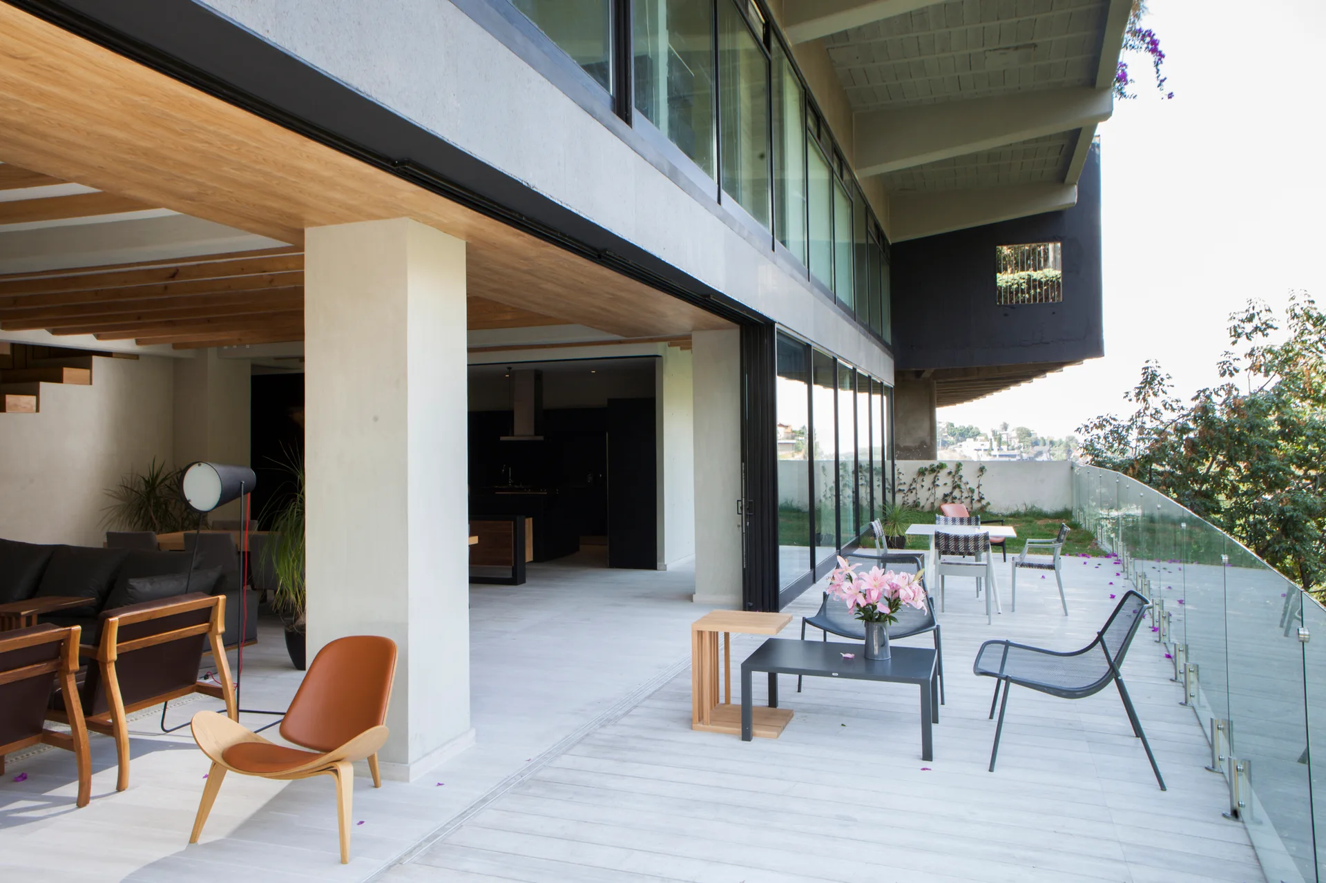 A modern outdoor terrace with black metal furniture and a glass railing connects to an indoor living area through large sliding glass doors.