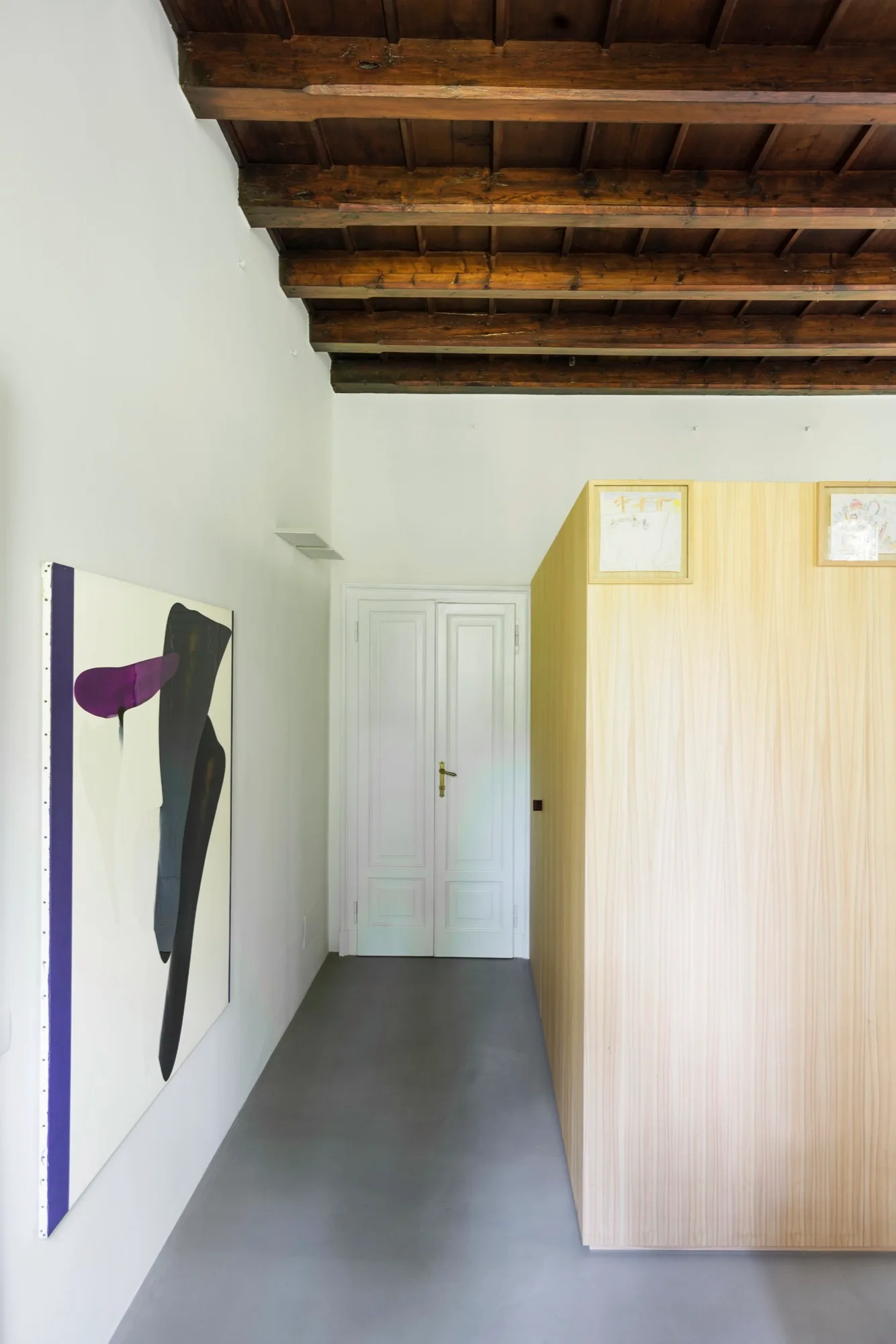 A modern hallway features a light wood partition wall, white double doors, grey flooring, and dark exposed wooden ceiling beams.
