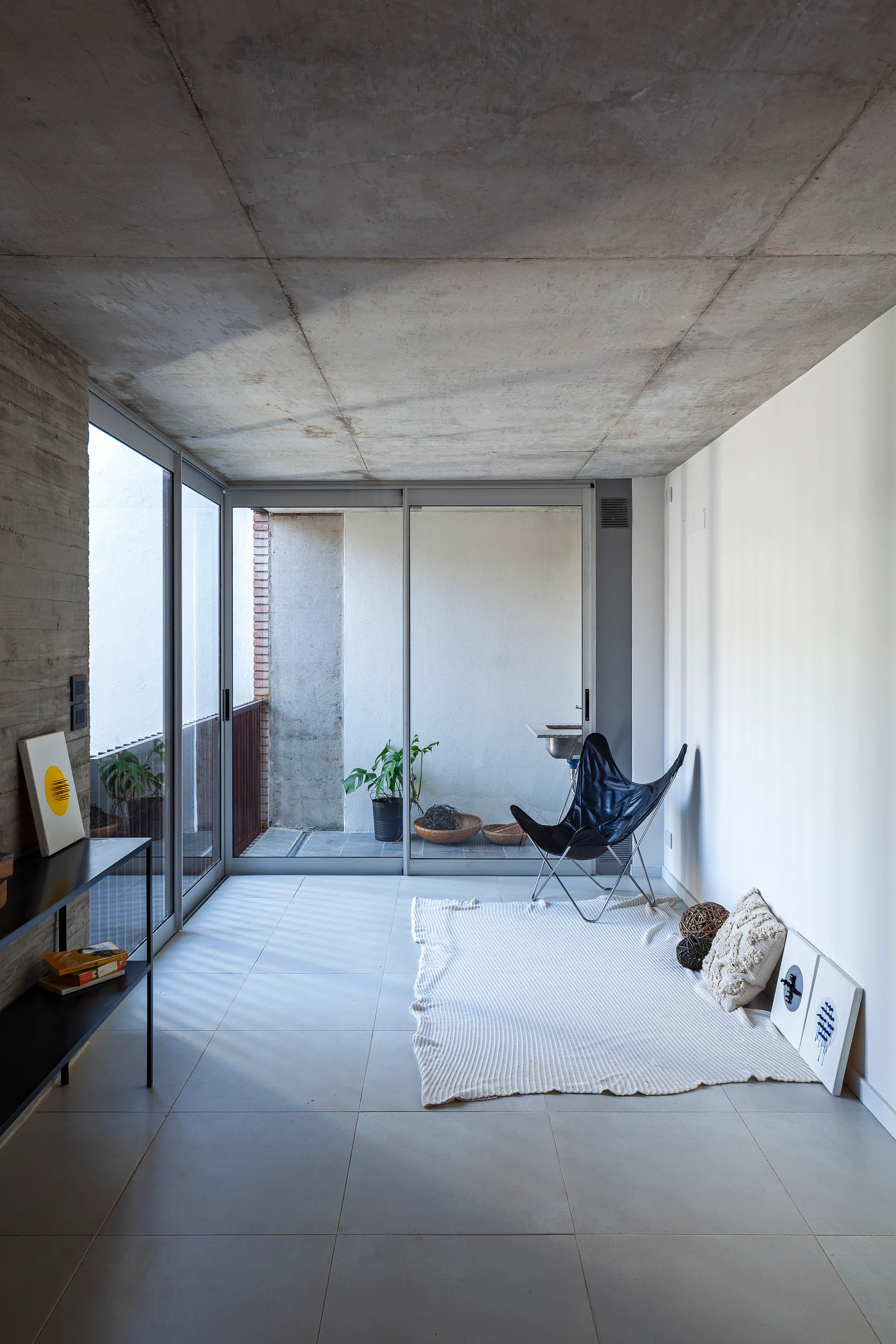 A minimalist living space featuring a raw concrete ceiling, tiled floors, a black butterfly chair, and sliding glass doors that open to a balcony.
