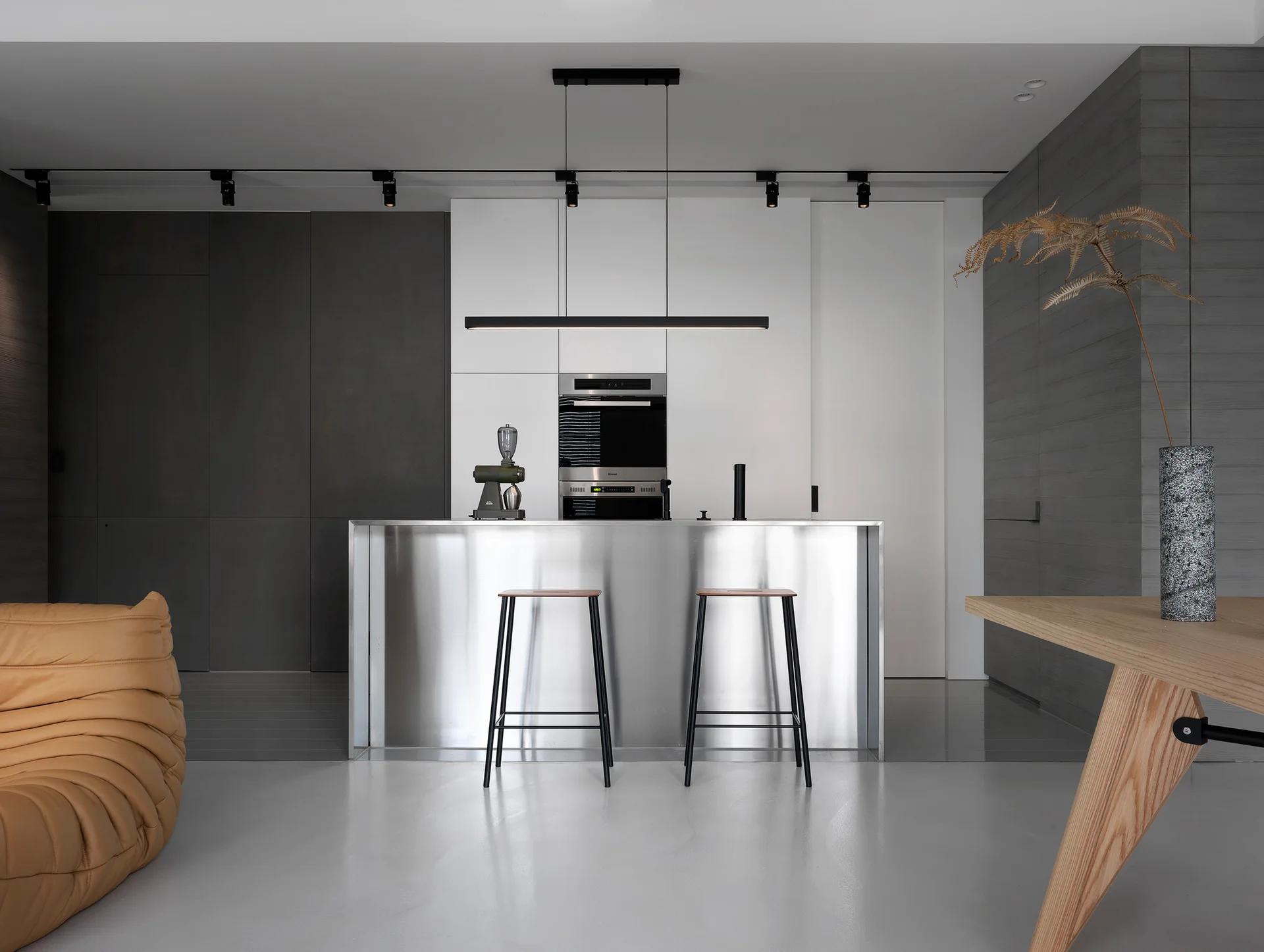 A minimalist kitchen features a central stainless steel island with bar stools, a linear pendant light, and built-in appliances set against grey wall panels.
