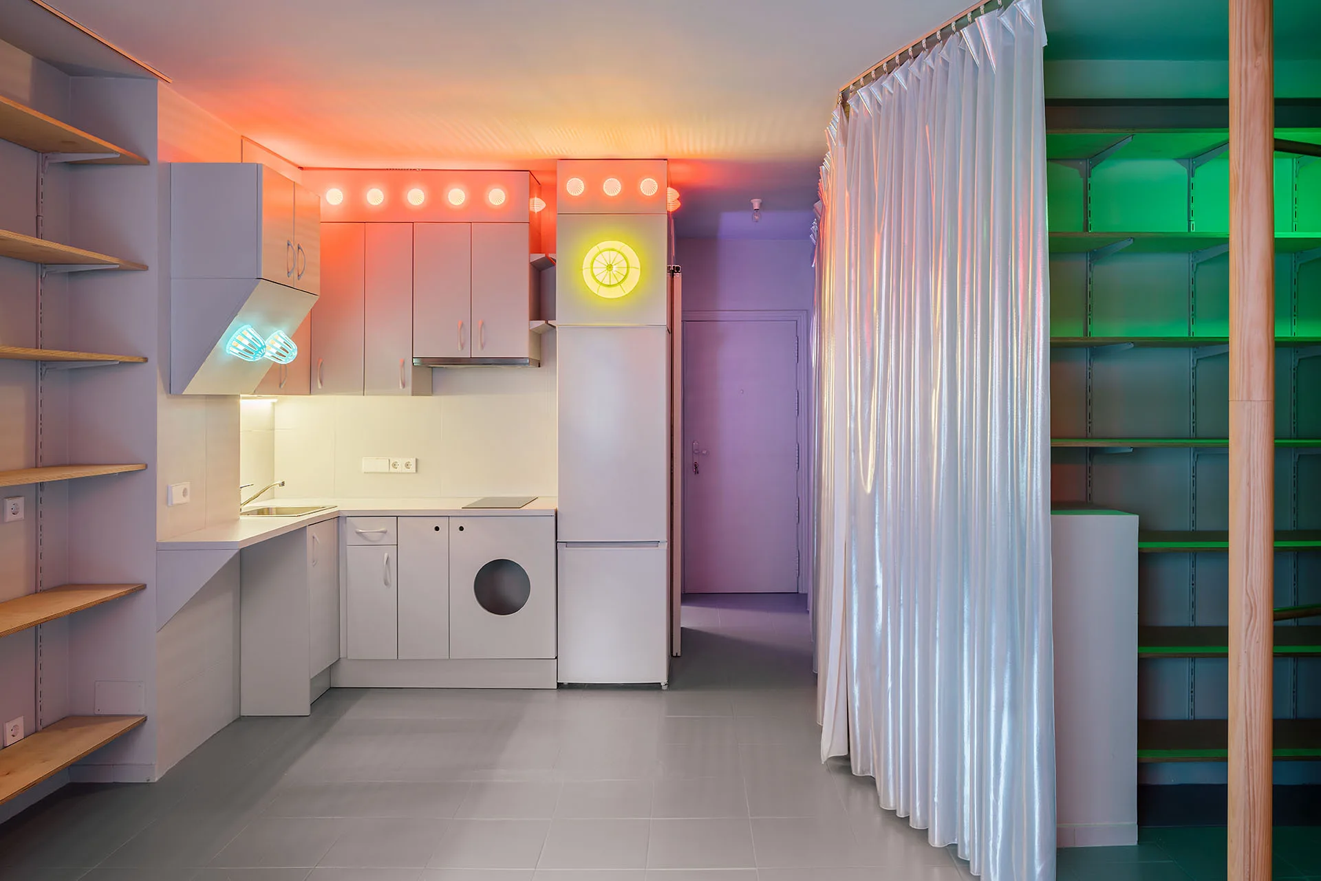 A modern kitchen with modular white cabinets, integrated washer, open wood shelves, a shiny room-dividing curtain, and multi-colored ambient neon lights.