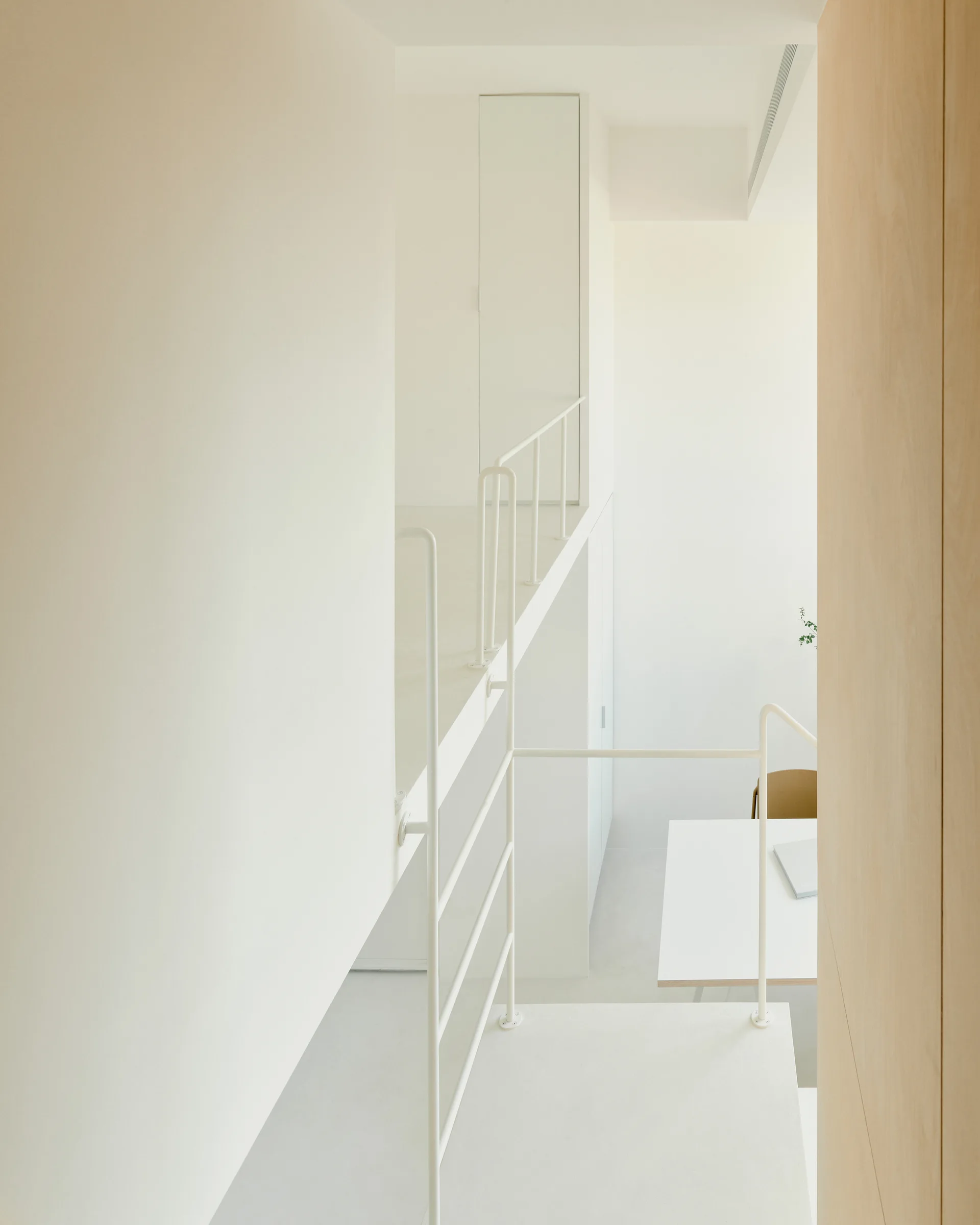 A minimalist interior featuring a white staircase with slender white tubular metal railings, matte white walls, and a landing with a flush door.