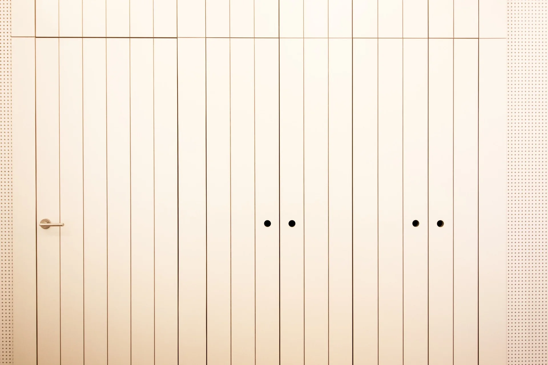 Minimalist off-white vertical paneling featuring an integrated flush door with a metal handle alongside cabinets with circular finger pull cutouts.