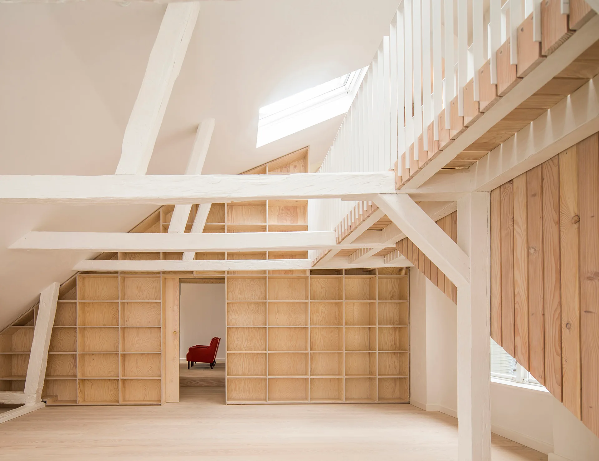 A bright attic space features custom light wood plywood shelving beneath sloped white ceilings with exposed timber beams.