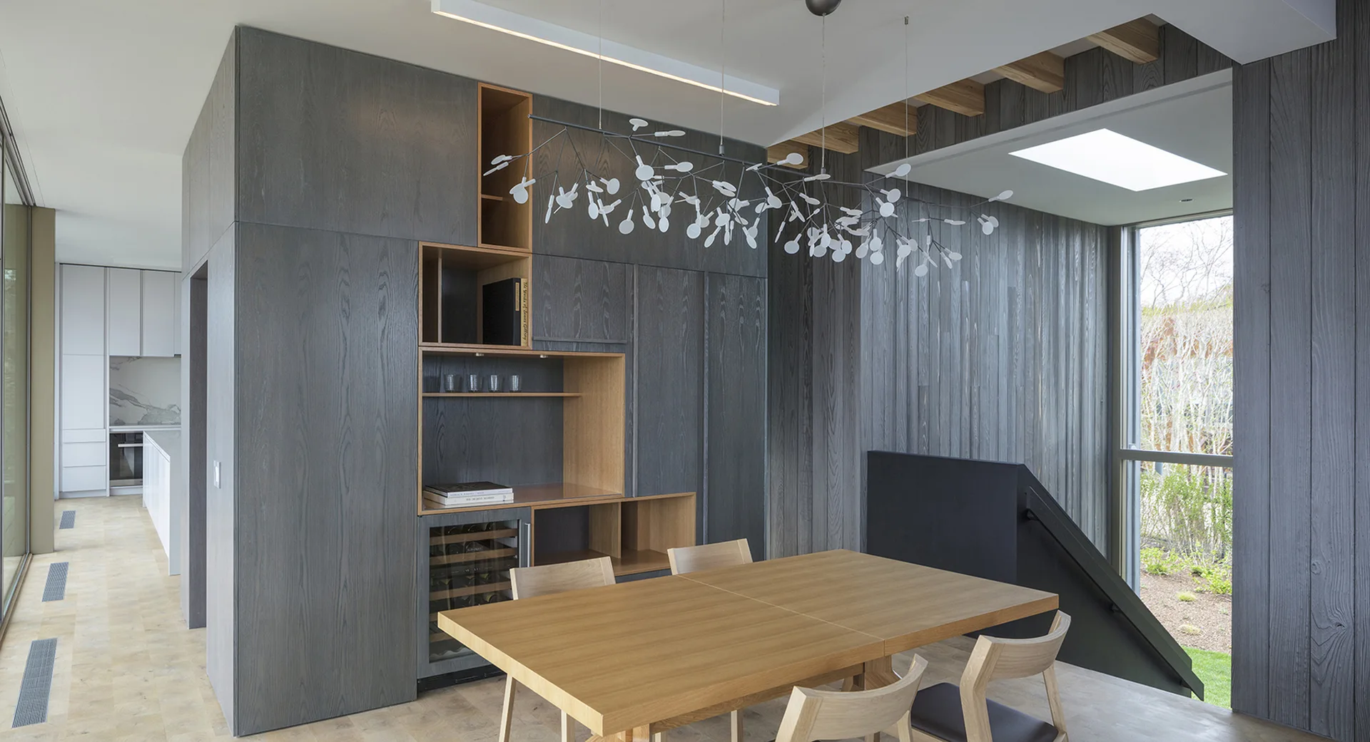 A modern dining room featuring a light wood table, charcoal gray paneled storage walls, an integrated wine fridge, and a branch-like chandelier.
