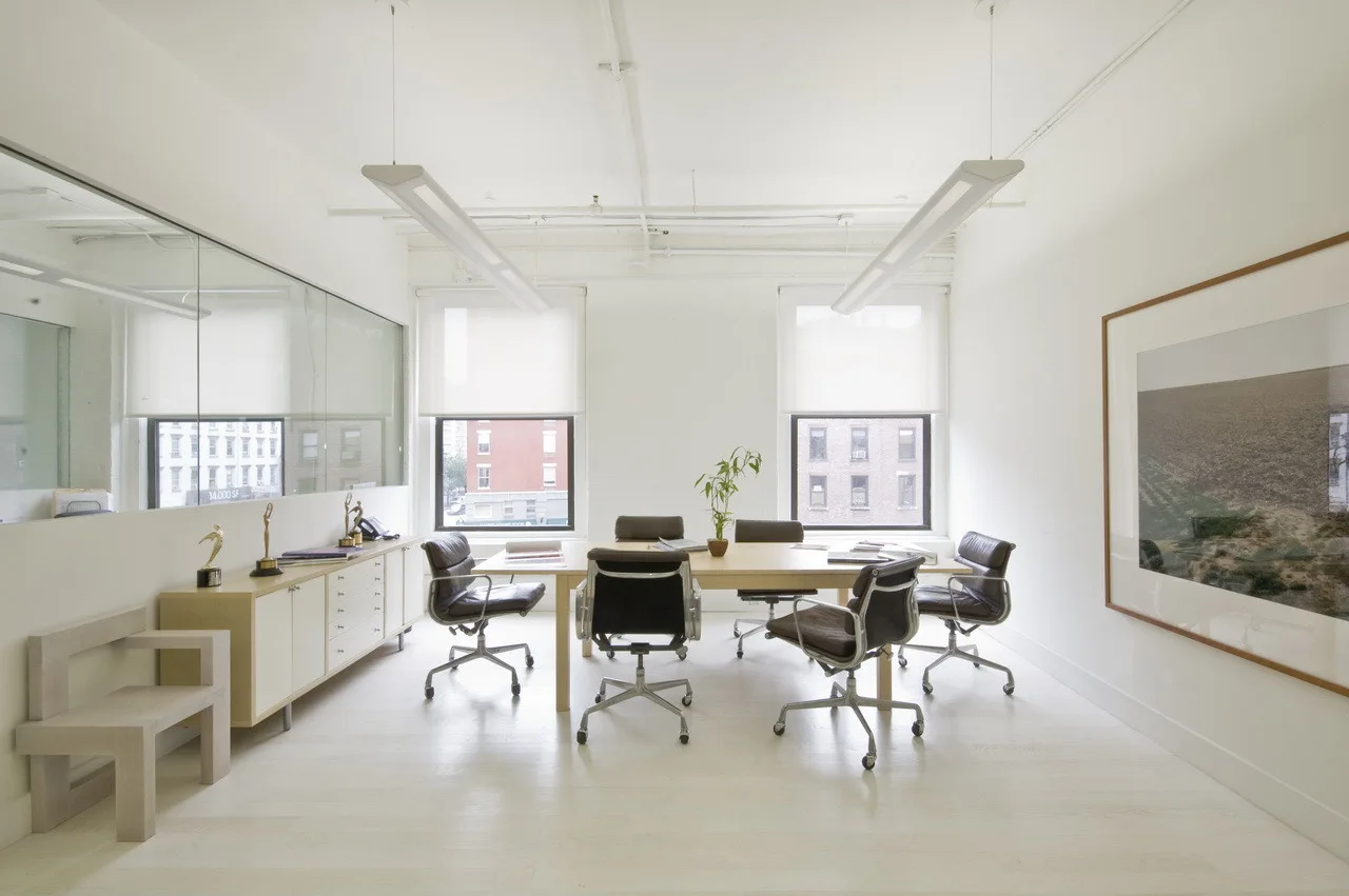 A minimalist conference room features a light wood table, black leather chairs, a long wall mirror, and two windows with white roller shades.