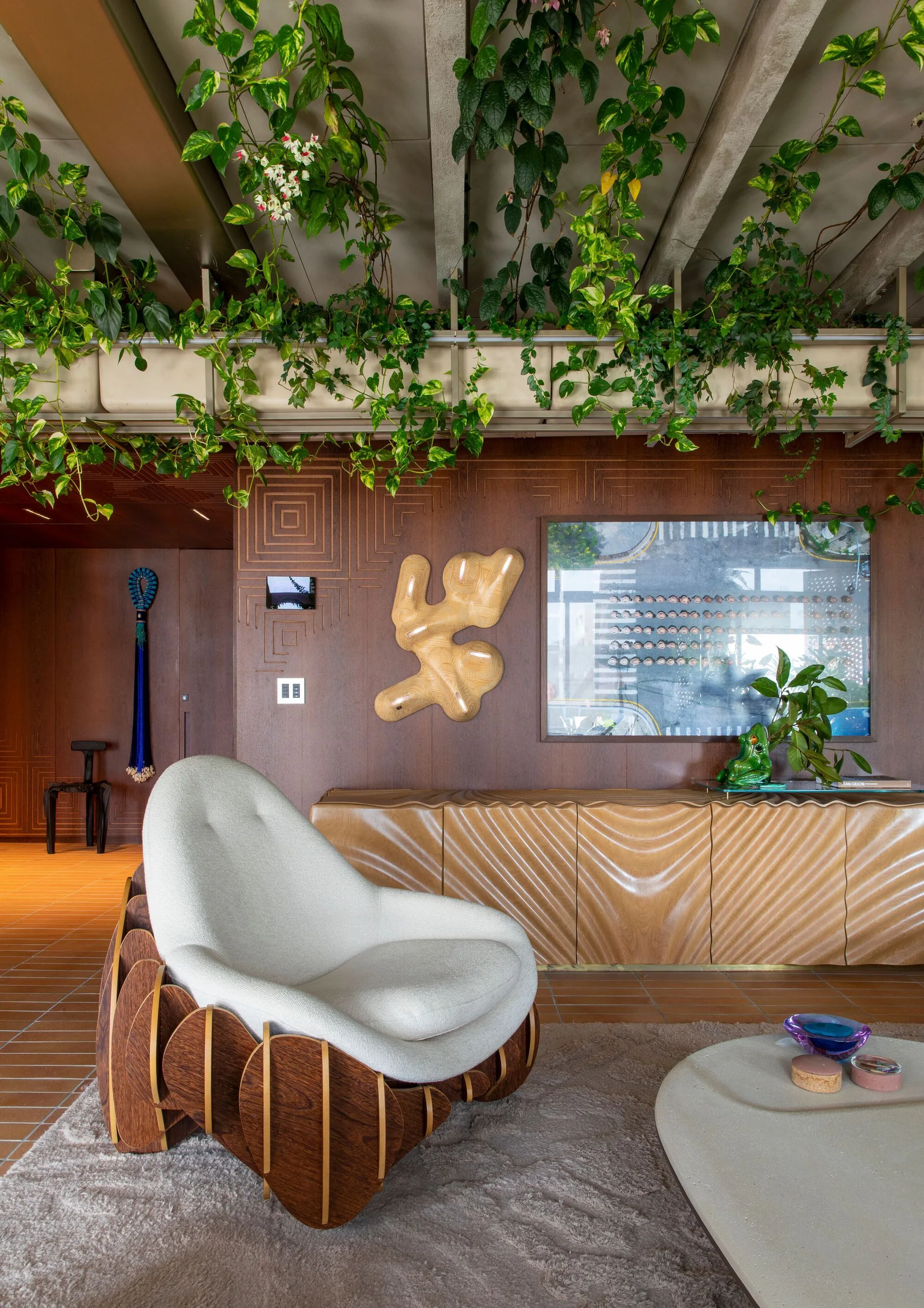 An organic modern living room featuring a sculptural cream armchair, a wavy wood sideboard, and green plants hanging from concrete ceiling beams.
