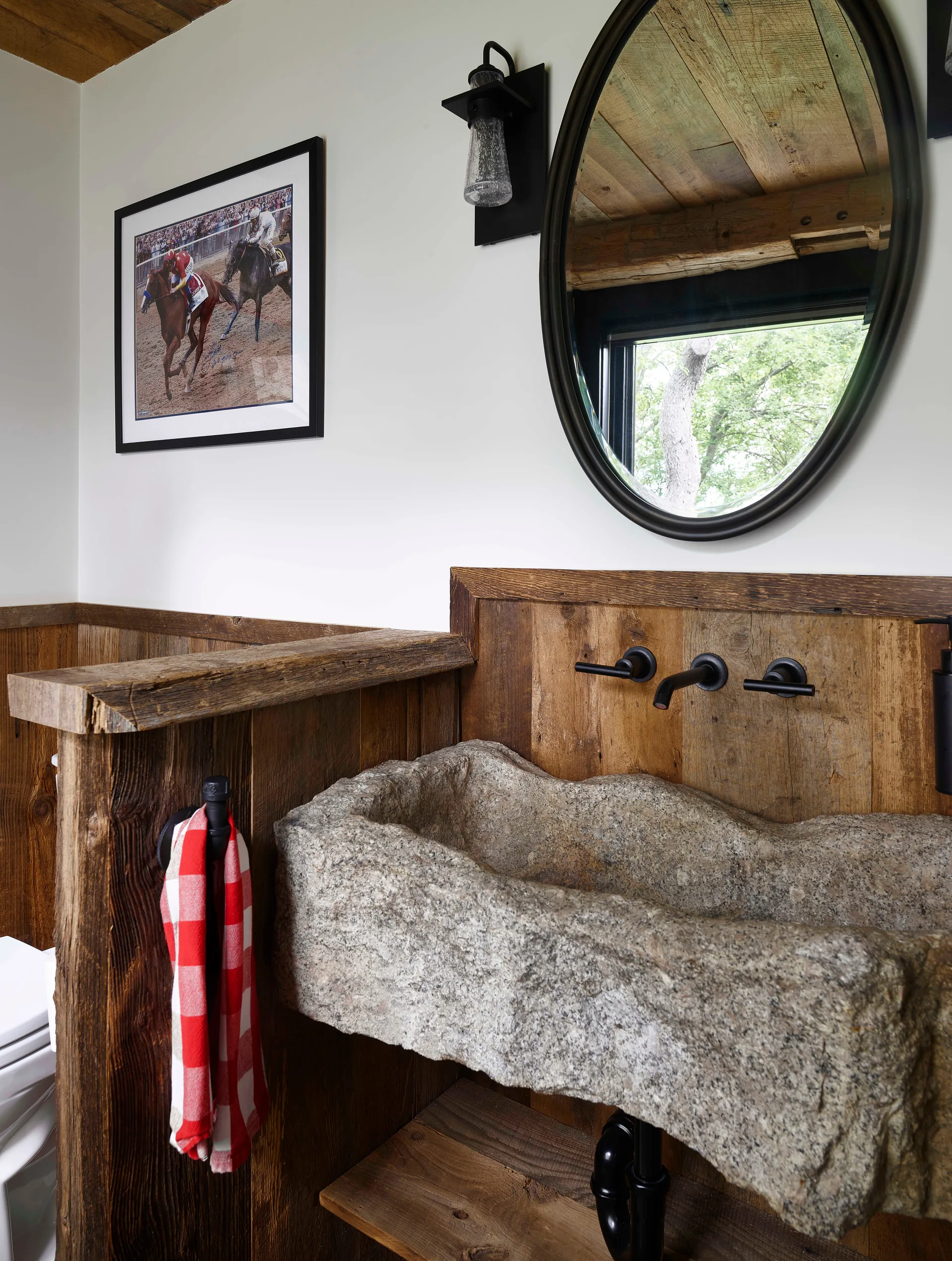 A rustic powder room featuring a rough stone basin sink, reclaimed wood wall paneling, and an oval black mirror.