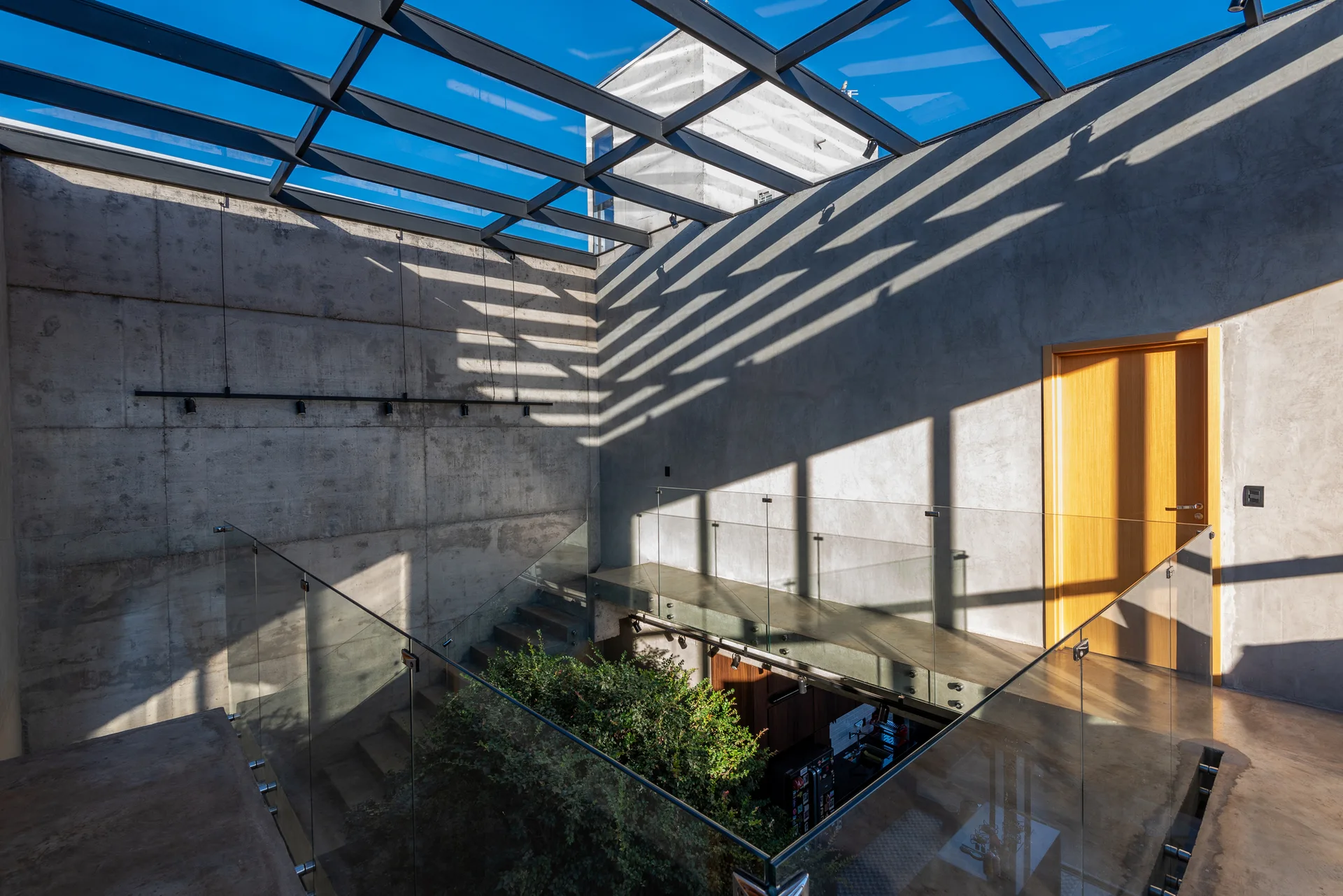 A minimalist staircase area with raw concrete walls, frameless glass railings, and a grid-patterned skylight roof casting geometric shadows.