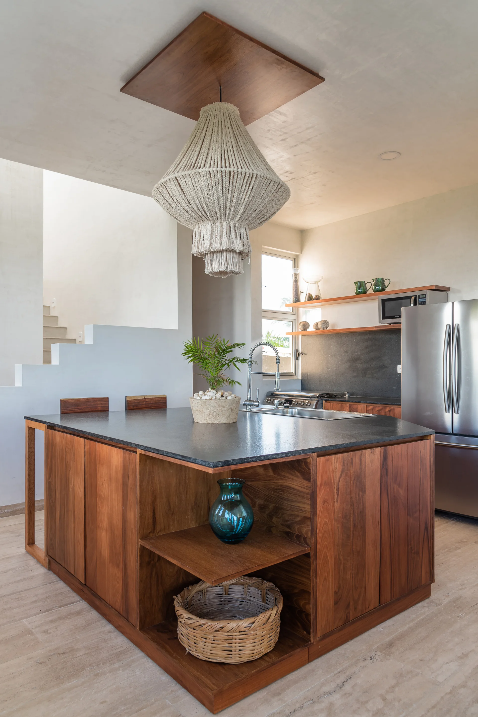 A modern kitchen features a warm wood island with a dark countertop, a large woven pendant light, and integrated open corner shelving.