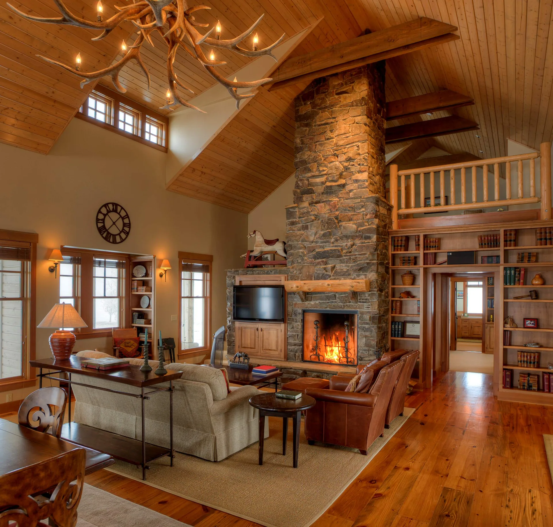 A rustic living room features a tall stone fireplace, a vaulted wood ceiling with an antler chandelier, and built-in bookshelves flanking a doorway.