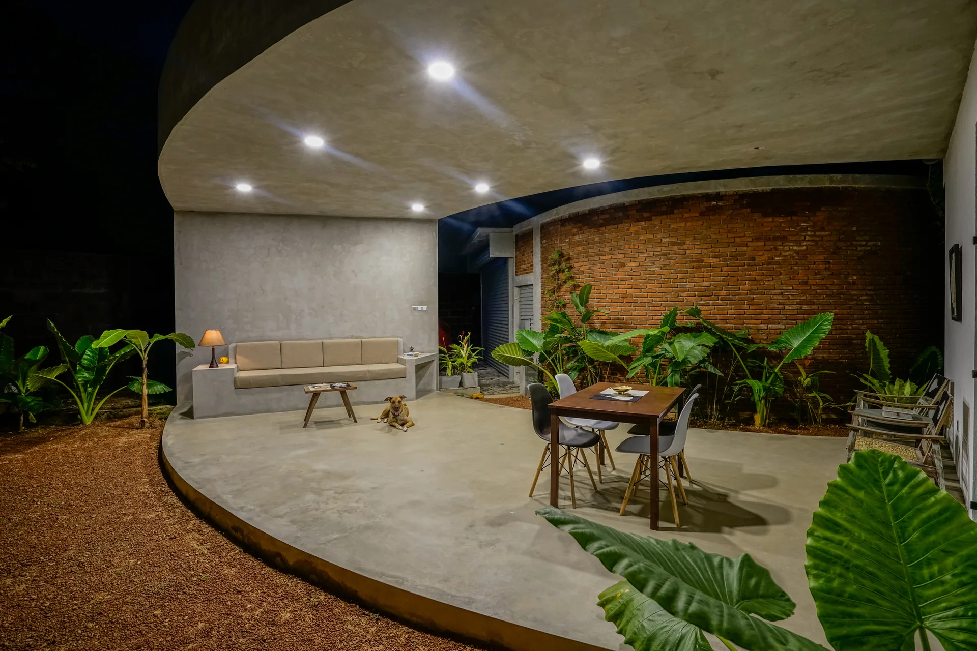 A modern covered patio at night featuring a curved concrete ceiling, a built-in sofa, a dining table, and a curved brick wall with lush plants.