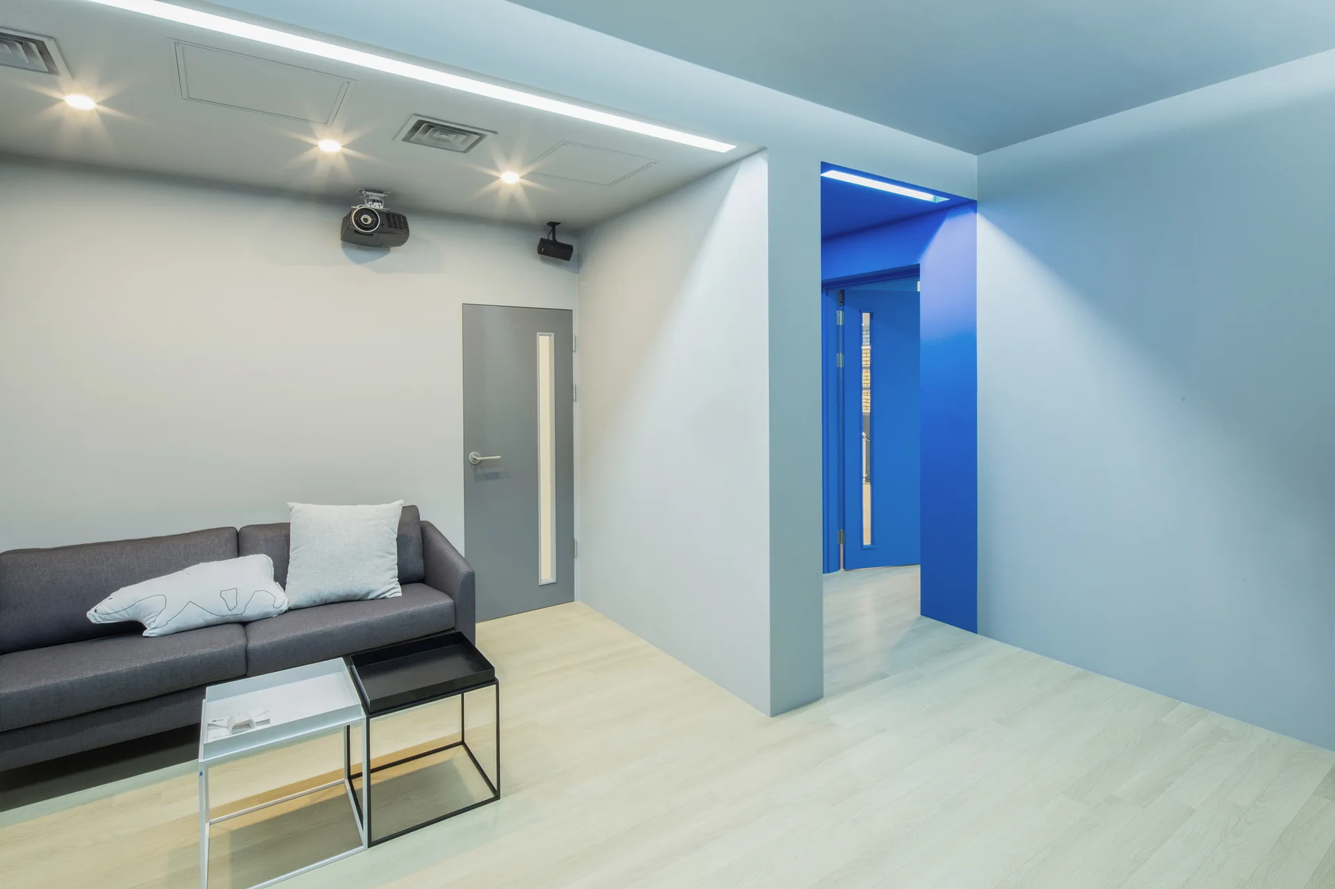 A minimalist lounge area features a grey sofa, light wood flooring, and a bright blue doorway passage under recessed ceiling lights.