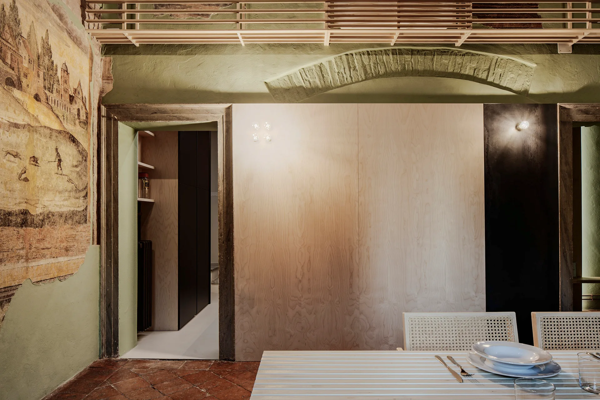 A dining room features a historic wall mural, sage green painted walls, raw plywood paneling, and a white dining table on terracotta tile.