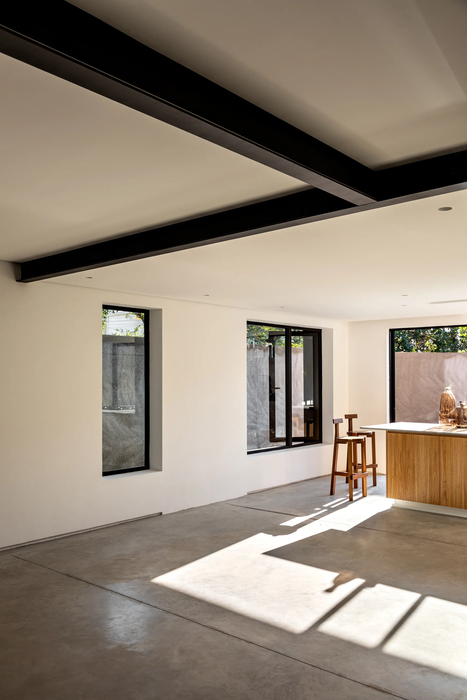 A minimalist kitchen features polished concrete floors, black ceiling beams, white walls, and a wooden island with bar stools.