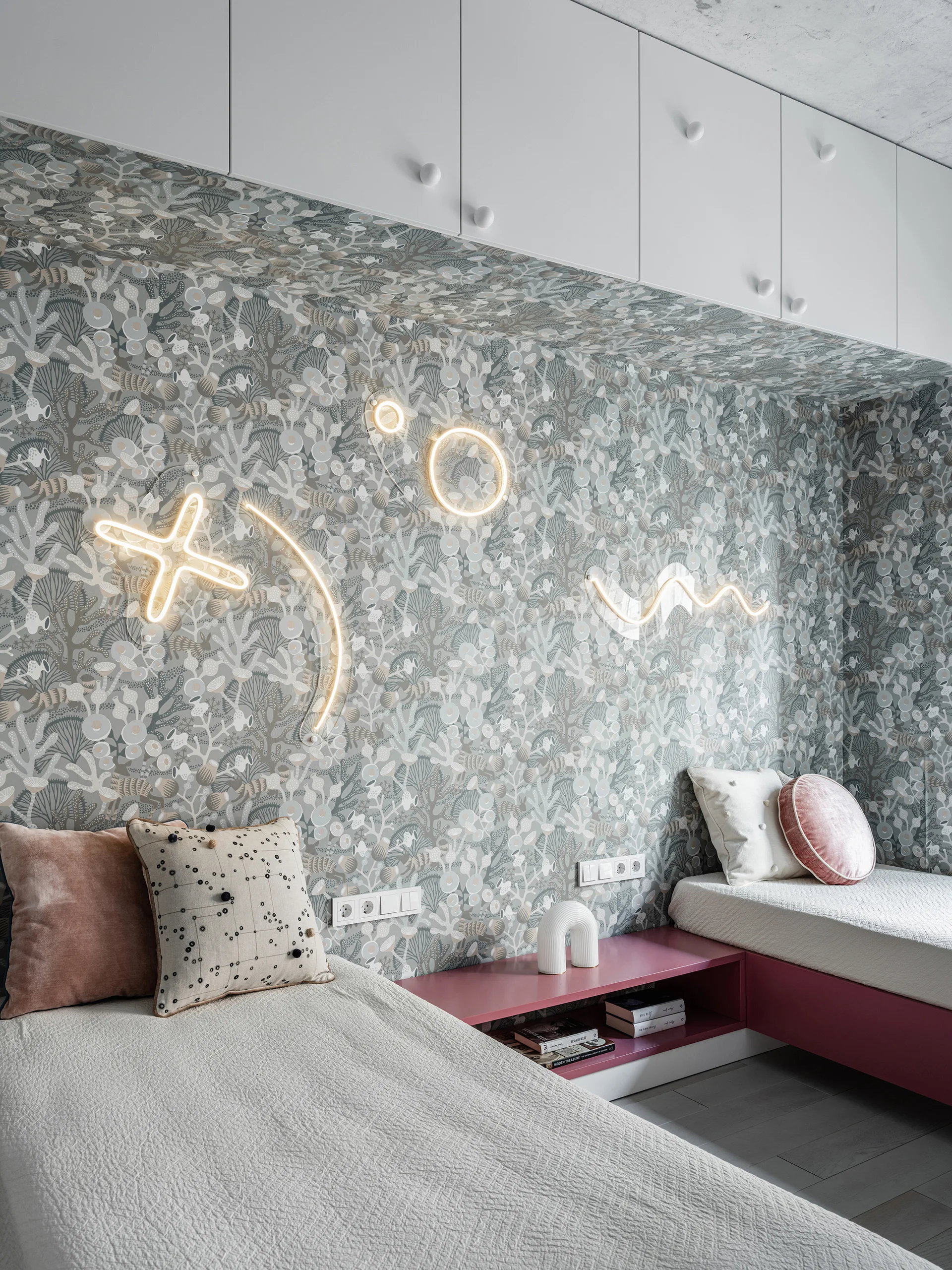 A shared kids' bedroom features two beds with textured white coverlets, botanical wallpaper, custom pink built-in shelving, and neon wall art.