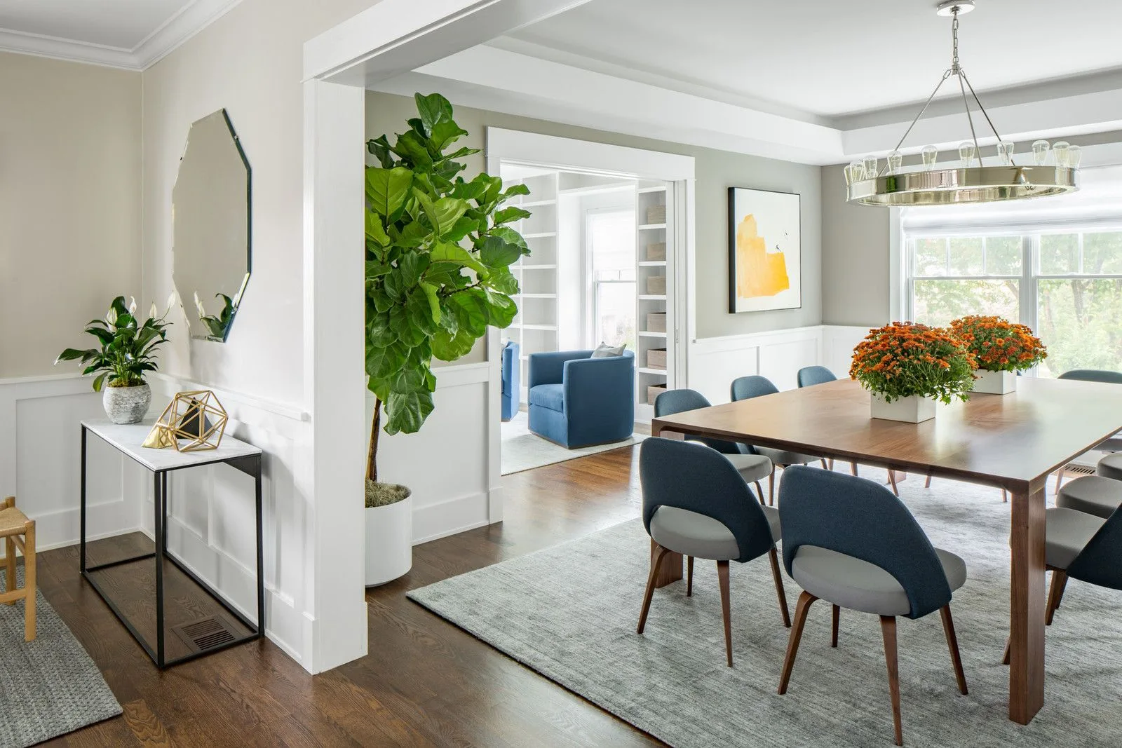 A bright dining room features a dark wood table, two-tone blue and grey chairs, a grey rug, wainscoting, and a view into an adjacent library room.