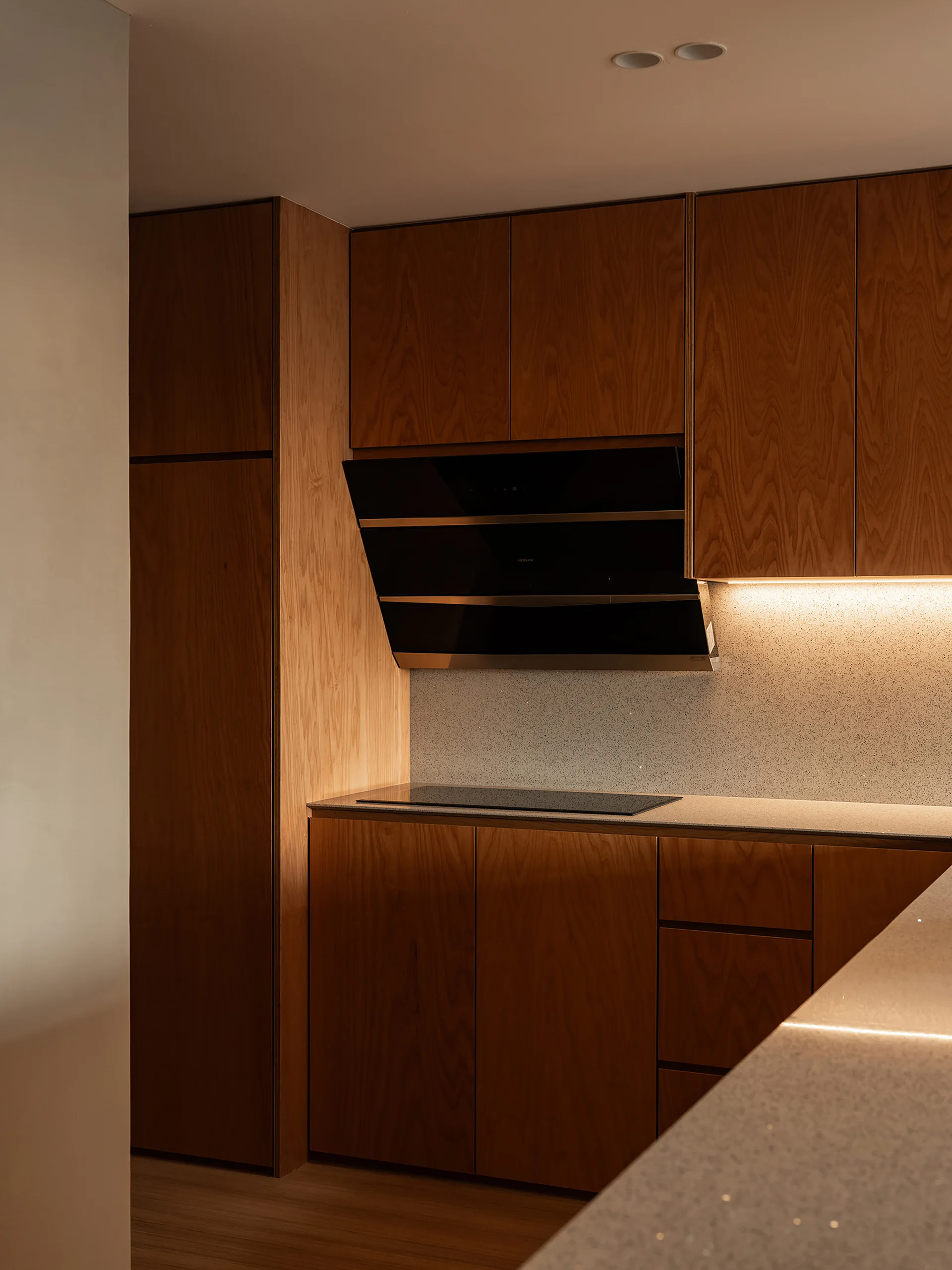 A modern kitchen featuring flat-panel wood cabinets, a black glass range hood, speckled countertops, and warm under-cabinet lighting.